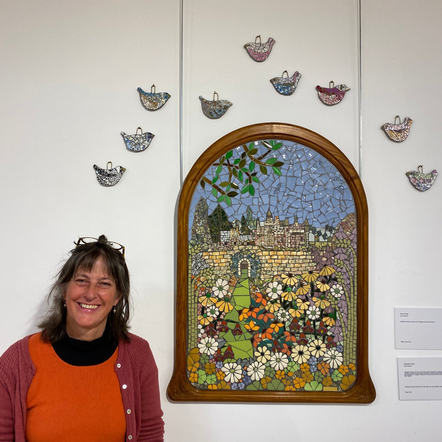 showing the mosaic on a white wall with mosaic birds above, and the artist beside it