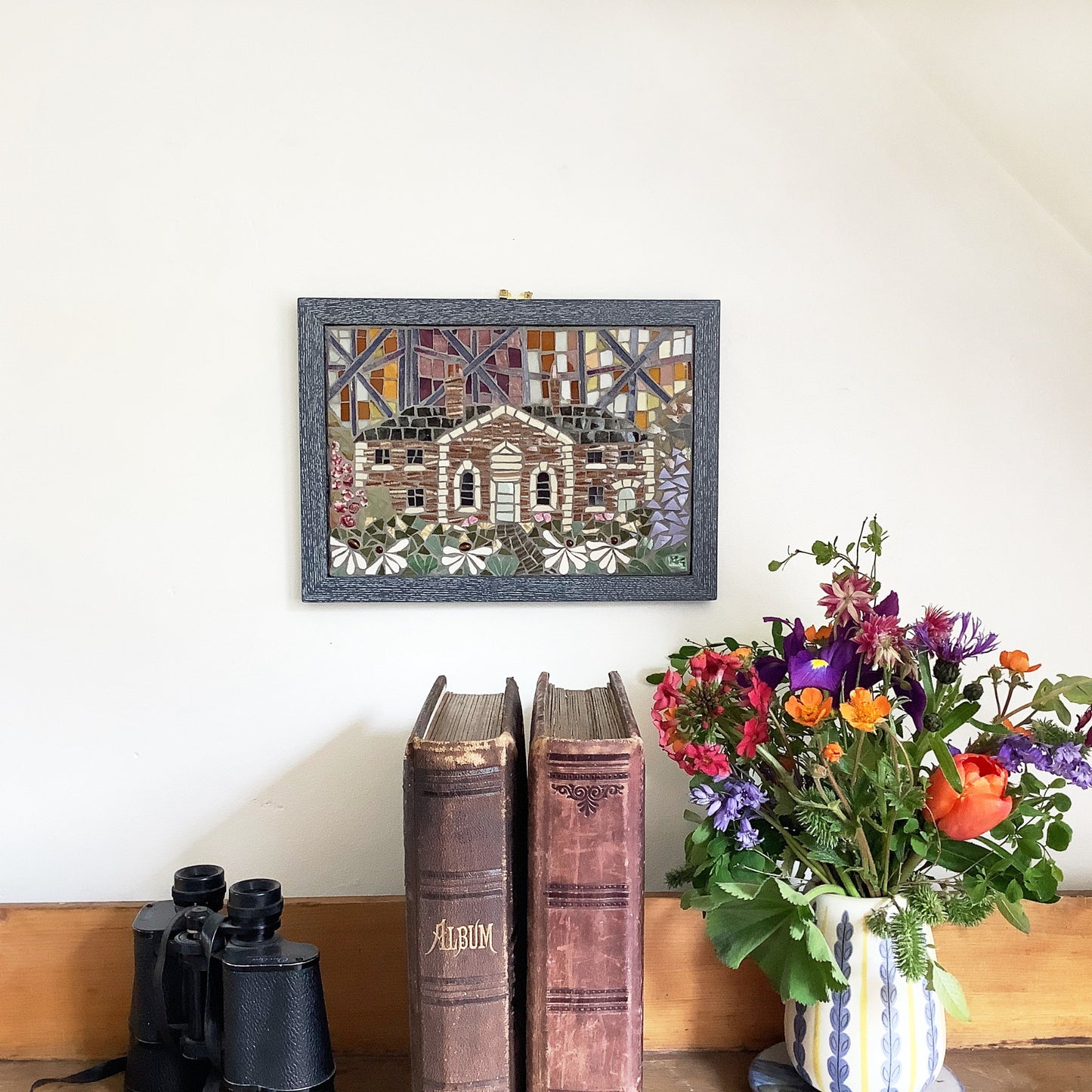 a mosaic picture of a cottage on a white wall, with books and flowers beside it
