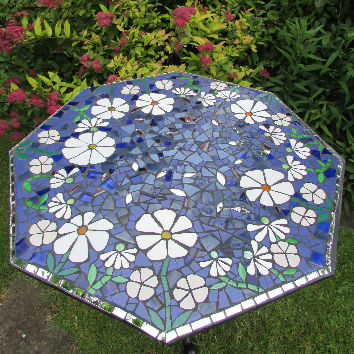 showing a hexagonal mosaic tabletop in a garden setting, with white flowers against a blue background. Mirror around the edges