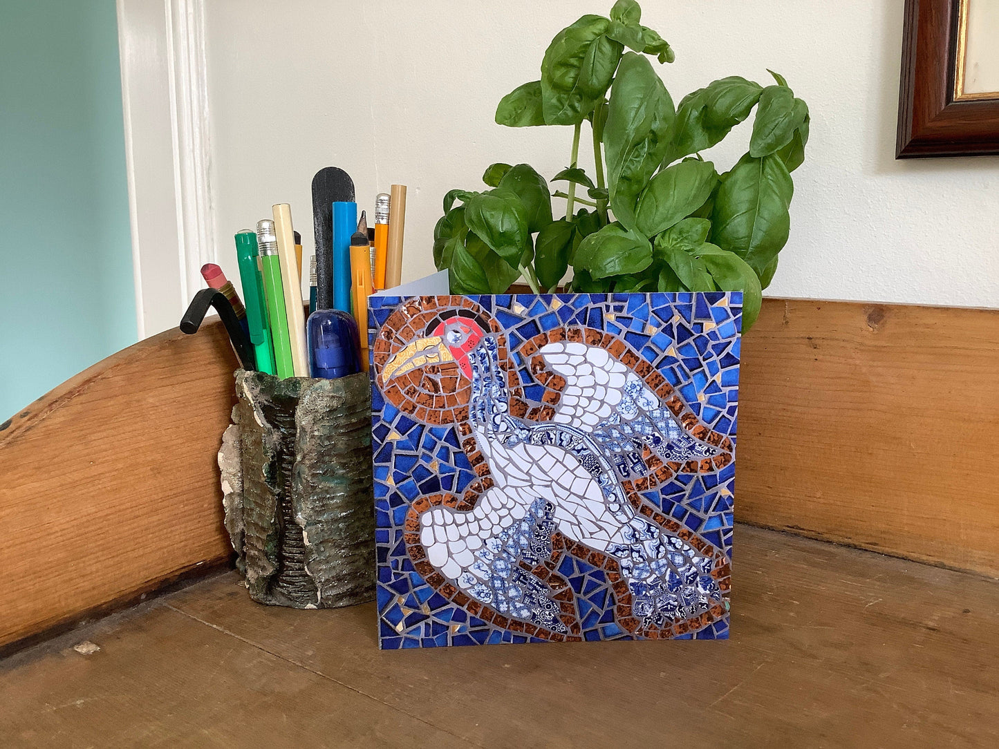 a square card with a crane bird with a halo depicted in mosaic. blue tile background. The bird has a white body and the tail & wing feathers are in blue and white traditional china. Photographed on display on a pine sideboard alongside a pot of pens and a plant
