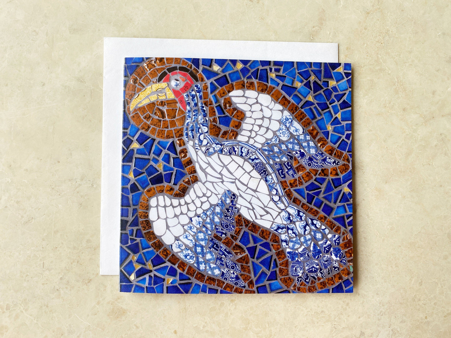 a square card with a crane bird with a halo depicted in mosaic. blue tile background. The bird has a white body and the tail & wing feathers are in blue and white traditional china. Photographed with a neutral background