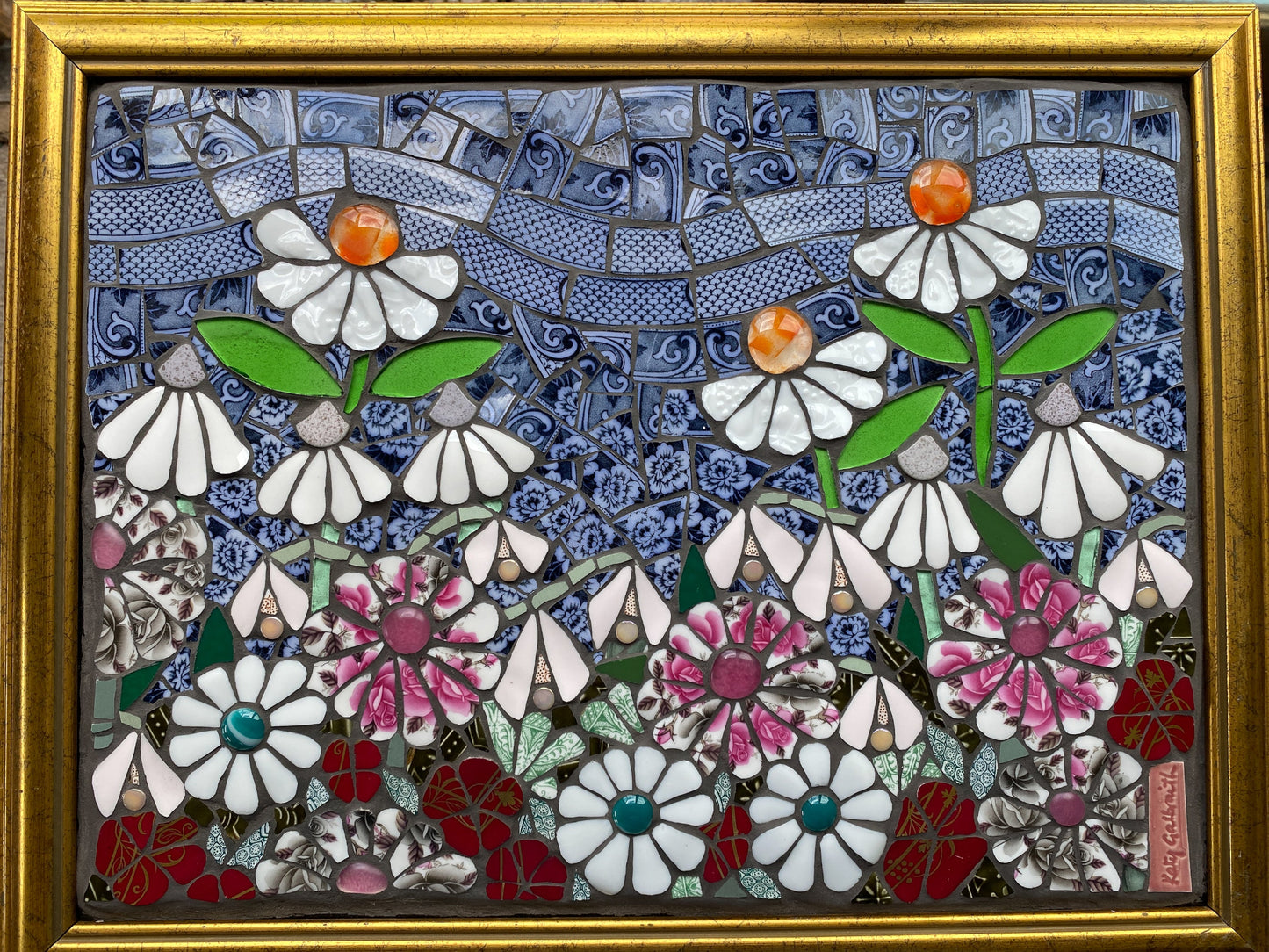 Mosaic artwork of flowers in a gold frame. White flowers with orange centres, pink and white flowers, blue sky made from crockery