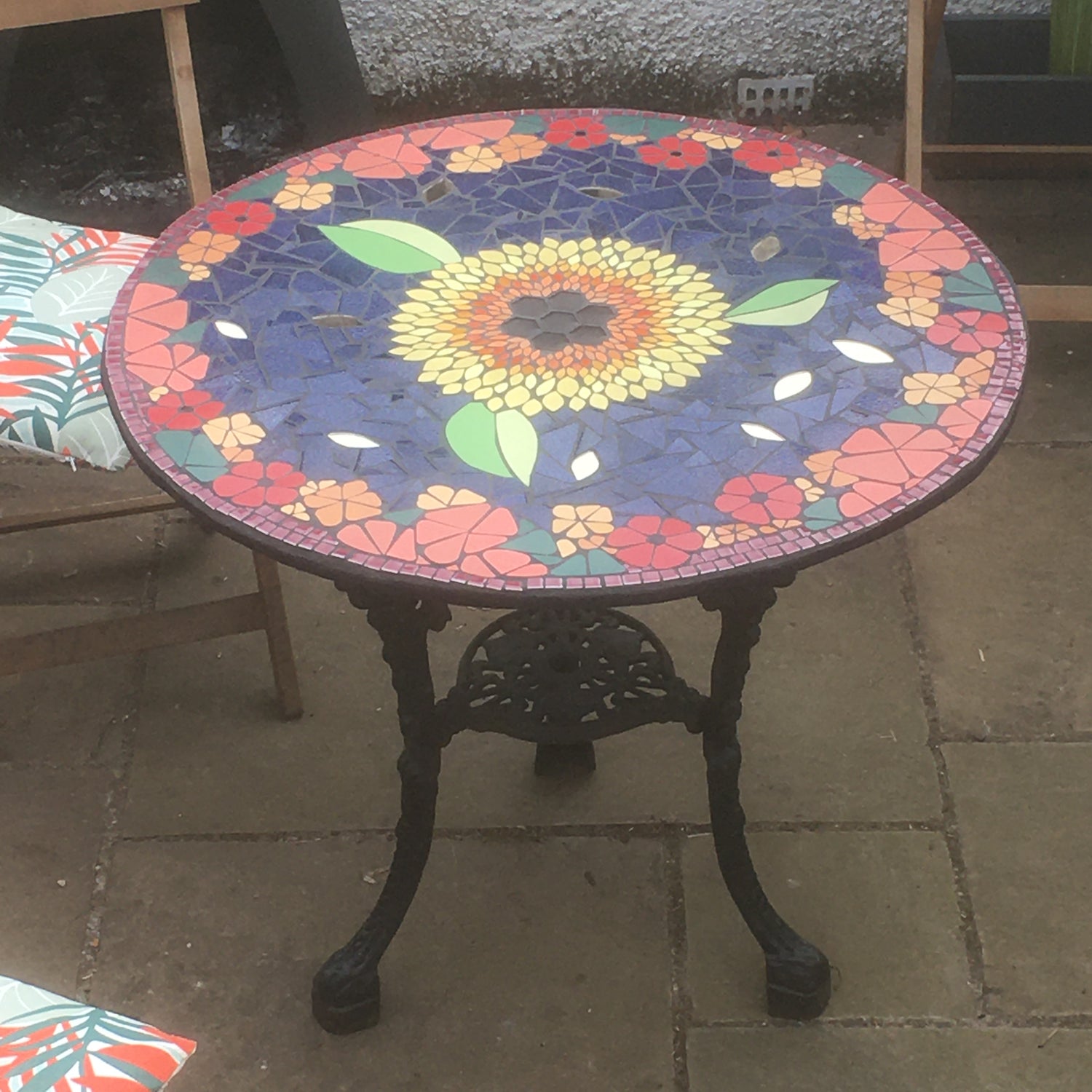 a circular mosaic garden table with a black base. the top is a mosaic of orange and red flowers with a yellow sunflower with leaves in the centre