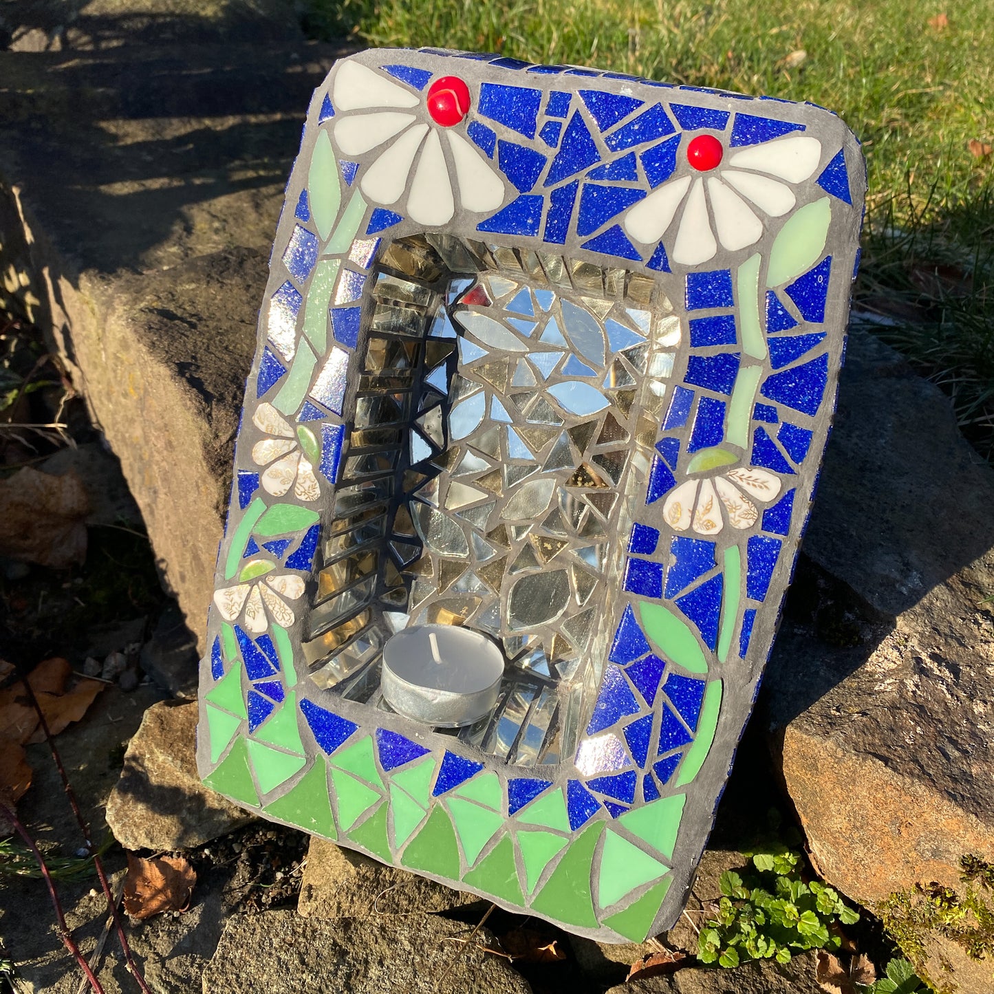 a small shrine in mosaic with two white daisies with red nuggets, against a blue background and green at the bottom. behind a candle is an area of mirror. product shown on a stone step with a bit of lawn visiible