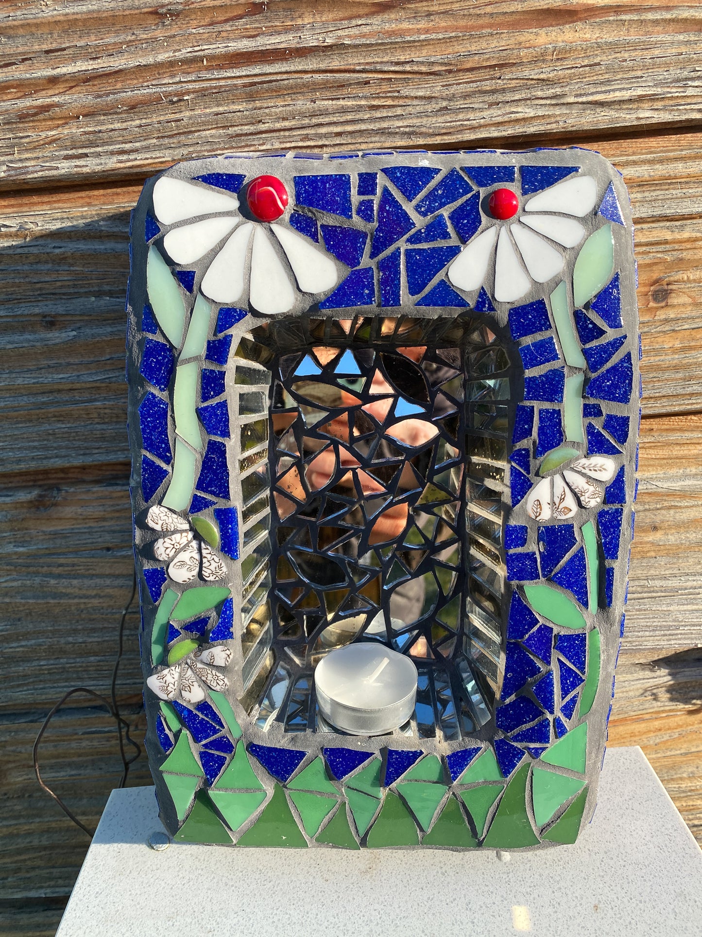 a small shrine in mosaic with two white daisies with red nuggets, against a blue background and green at the bottom. behind a candle is an area of mirror. product shown on a white base with wooden background