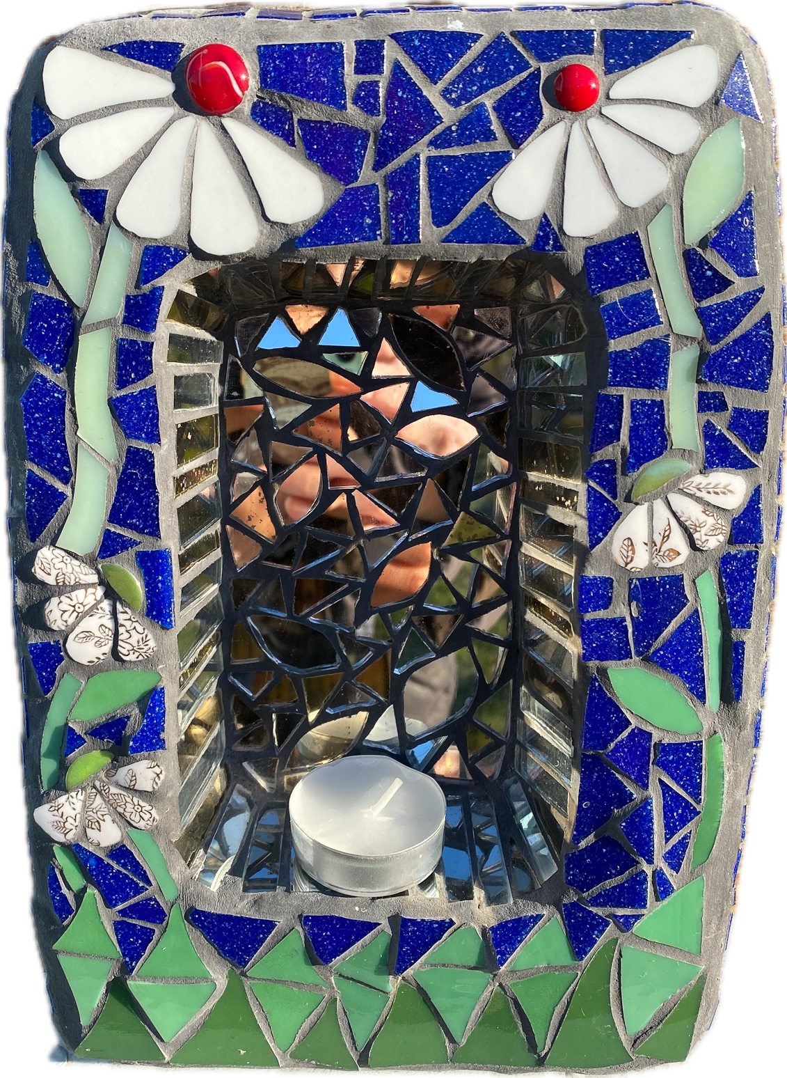 a small shrine in mosaic with two white daisies with red nuggets, against a blue background and green at the bottom. behind a candle is an area of mirror.