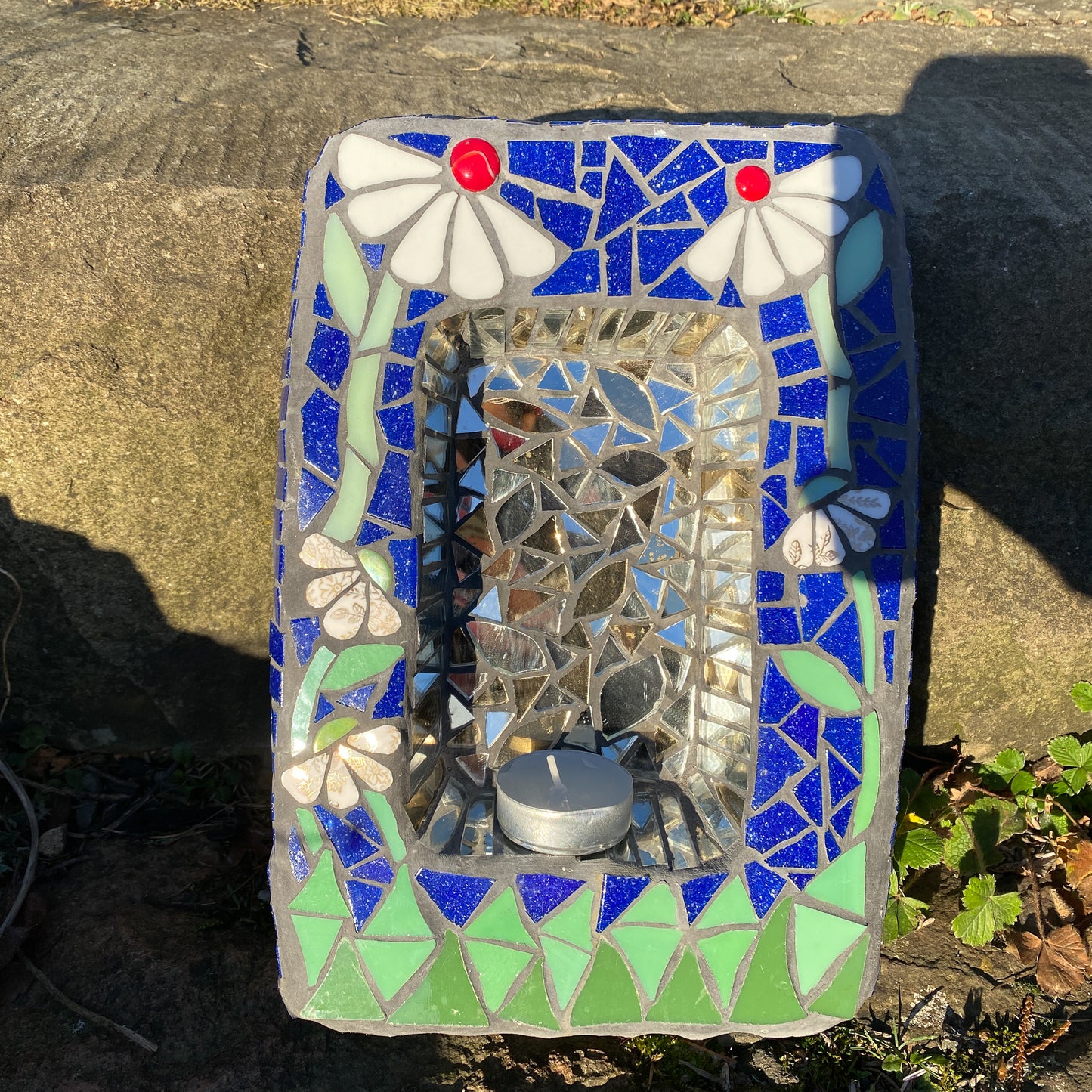 a small shrine in mosaic with two white daisies with red nuggets, against a blue background and green at the bottom. behind a candle is an area of mirror. product shown on a stone step