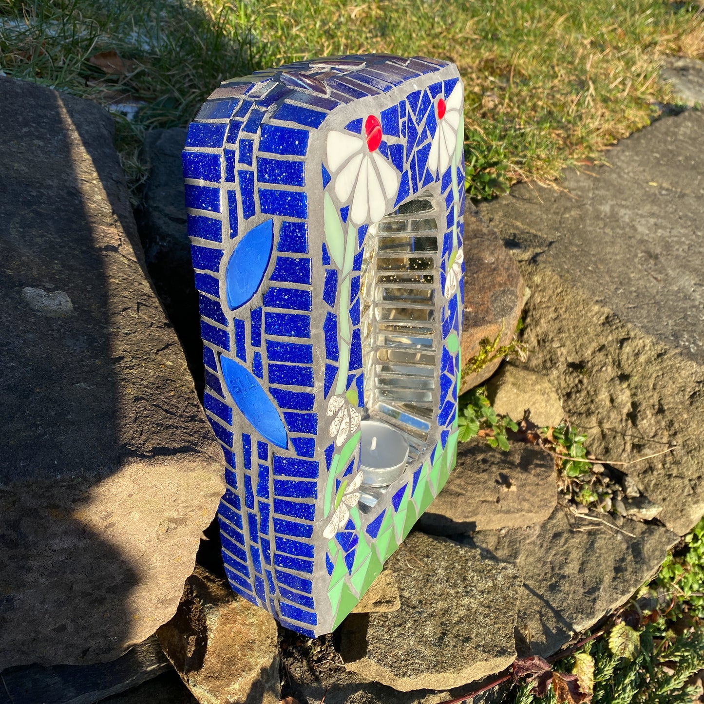 a small shrine in mosaic with two white daisies with red nuggets, against a blue background and green at the bottom. image showing the side view in blue mosaic. product shown on a step with grass