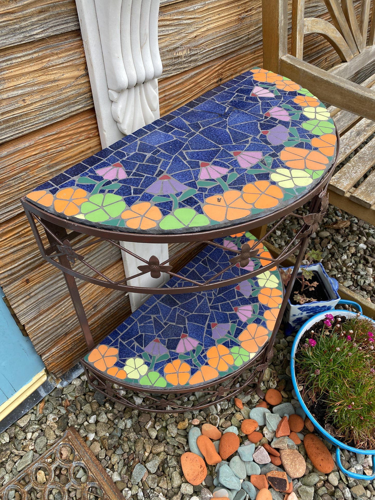 half moon patio table with mosaic surfaces. Two surfaces, with orange and lime green flowers edge the blue centre . The table is a metal vintage one painted dark brown. Photographed straight on and in an outdoors seating area