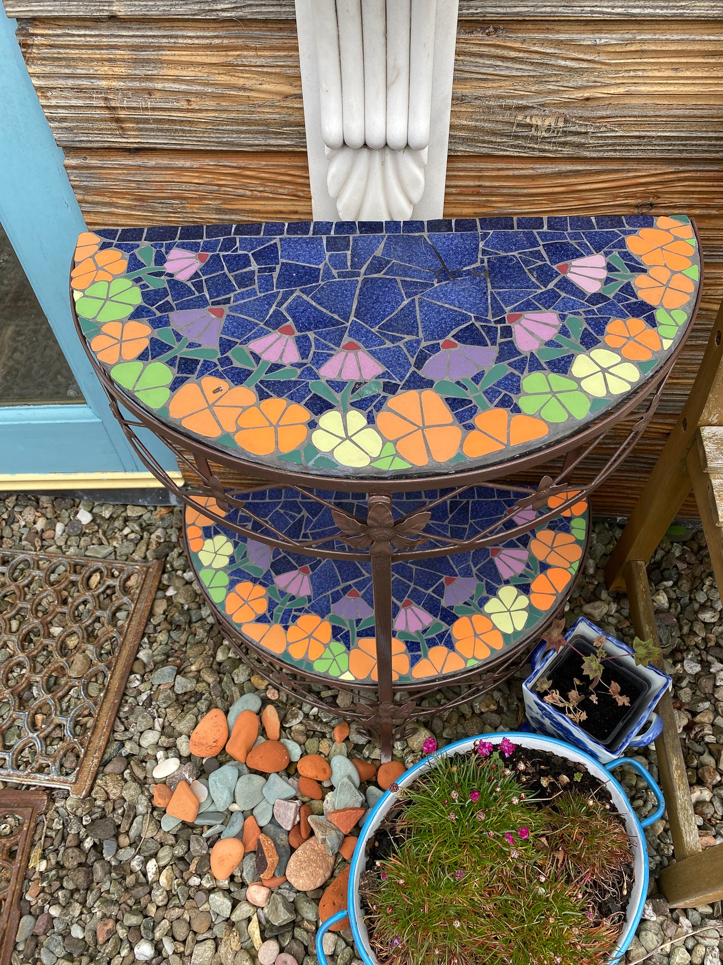 half moon patio table with mosaic surfaces. Two surfaces, with orange and lime green flowers edge the blue centre . The table is a metal vintage one painted dark brown. Photographed straight on and in an outdoors context