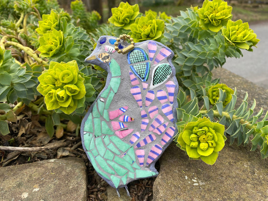 Mosaic peacock as viewed from the side rather than full feather display, with a green body, and pink and blue stripy tail feathers, with a pair of tennis racket heads in the tail.  Made from crockery. photographed with a foliage background