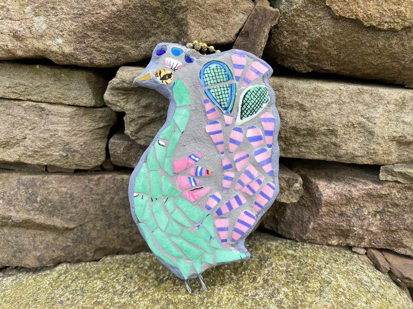 Mosaic peacock, viewed from the side. With a green body, and pink and blue stripy tail feathers, with a pair of tennis racket heads in the tail. Made from crockery. photographed with a drystone wall background