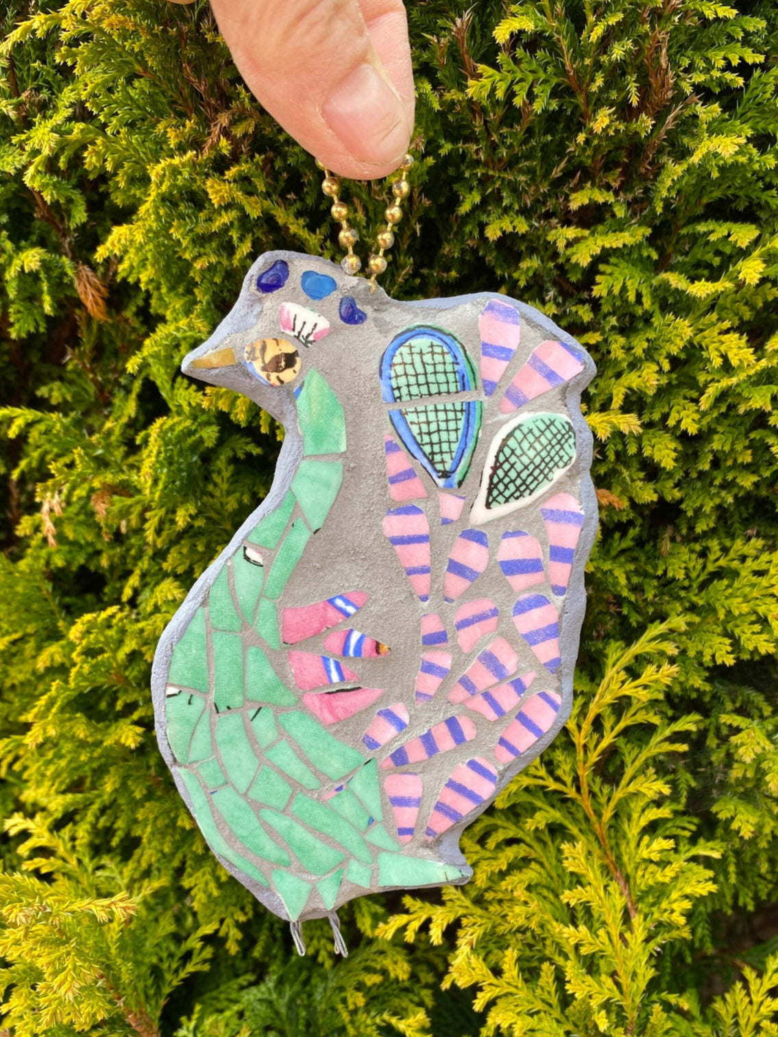 Mosaic peacock, viewed from the side. With a green body, and pink and blue stripy tail feathers, with a pair of tennis racket heads in the tail. Made from crockery. photographed held by the hanging mechanism with a foliage background