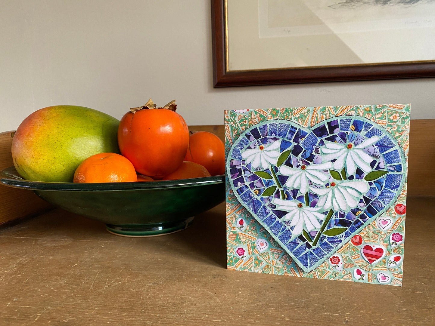 Square card using a mosaic image on the front. The main image is a heart shape with 5 white daisies inside it, and the background completing the square shape is a soft green with varying heart shapes, all in mosaic. The card is on display with a bowl of fruit beside it on a pine dresser