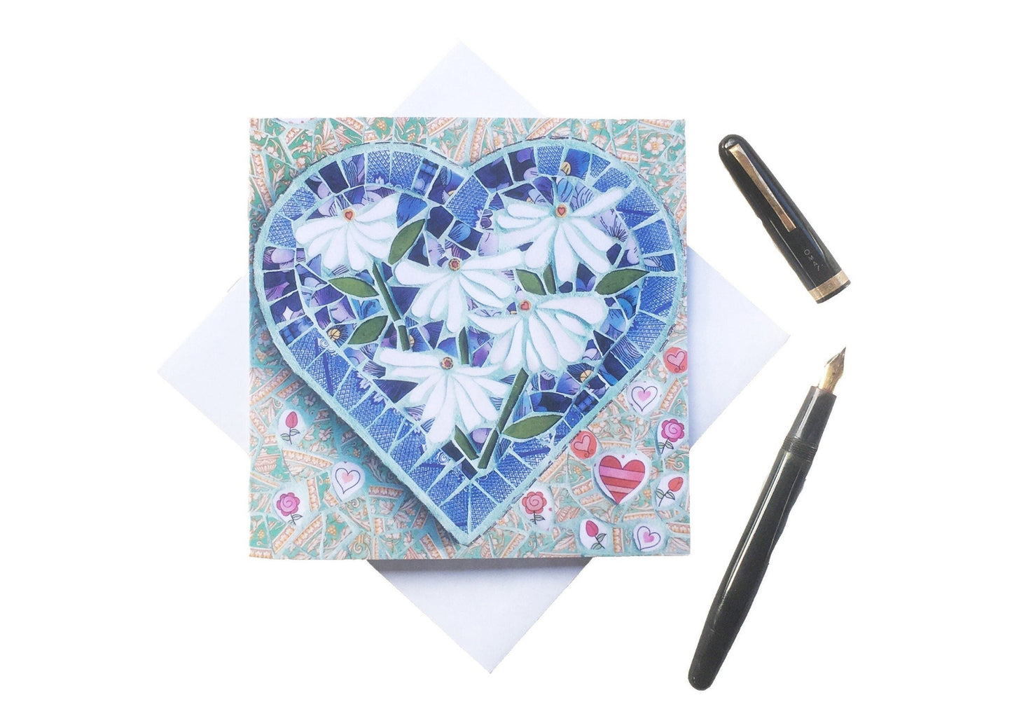 Square card using a mosaic image on the front. The main image is a heart shape with 5 white daisies inside it, and the background completing the square shape is a soft green with varying heart shapes, all in mosaic. The card is sitting on a square envelope, with a vintage ink pen beside it. Photographed on a white background.
