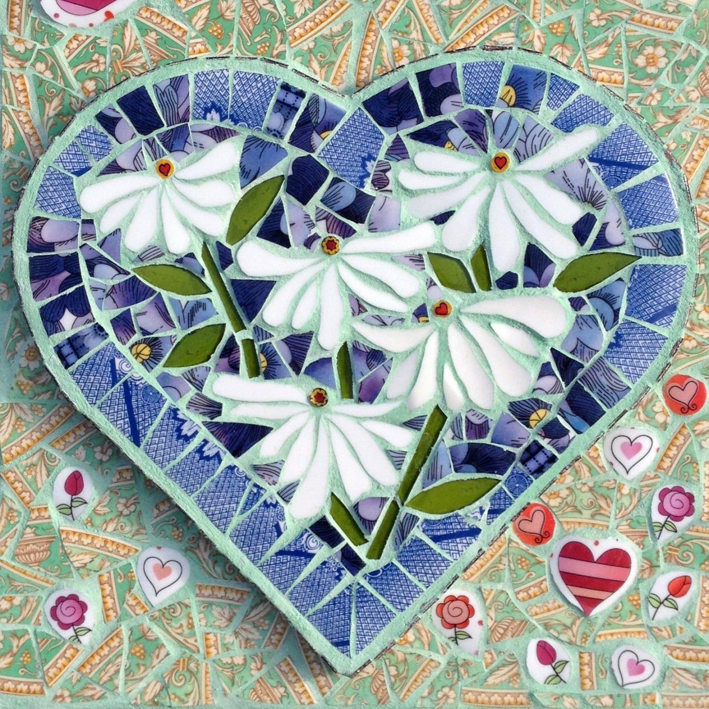 Square card using a mosaic image on the front. The main image is a heart shape with 5 white daisies inside it, and the background completing the square shape is a soft green with varying heart shapes, all in mosaic. The card is sitting on a square envelope, with a vintage ink pen beside it. Photographed on a white background.