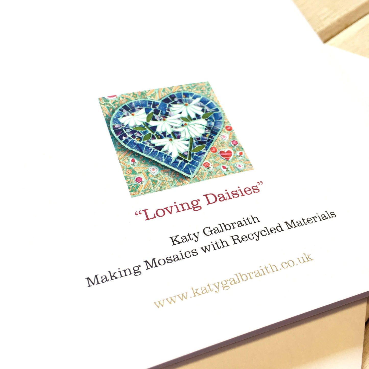 showing part of the reverse side of the card, with white card, small image of the card design and the artist details - Katy Galbraith, making mosaics with recycled materials and website www.katygalbraith.co.uk on it.