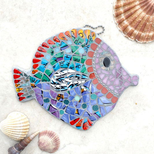 A mosaic fish made from bright purple and turquoise crockery, pink face surrounded by curved red tiles.  Fins and tails are in a red china.  Hangs by a ball chain at the top.  Photographed with shells.