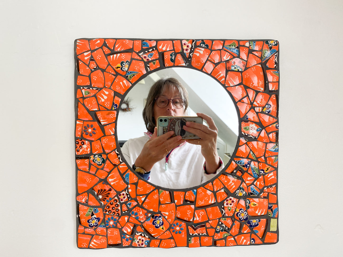 Bright orange mosaic mirror made from Turkish pottery. Square in shape with a circular mirror. You can see the artist taking the photo in the mirror