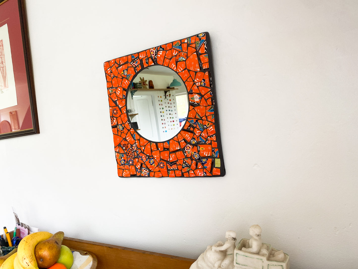 Bright orange mosaic mirror made from Turkish pottery. Square in shape with a circular mirror. Photographed from an angle showing it on a wall