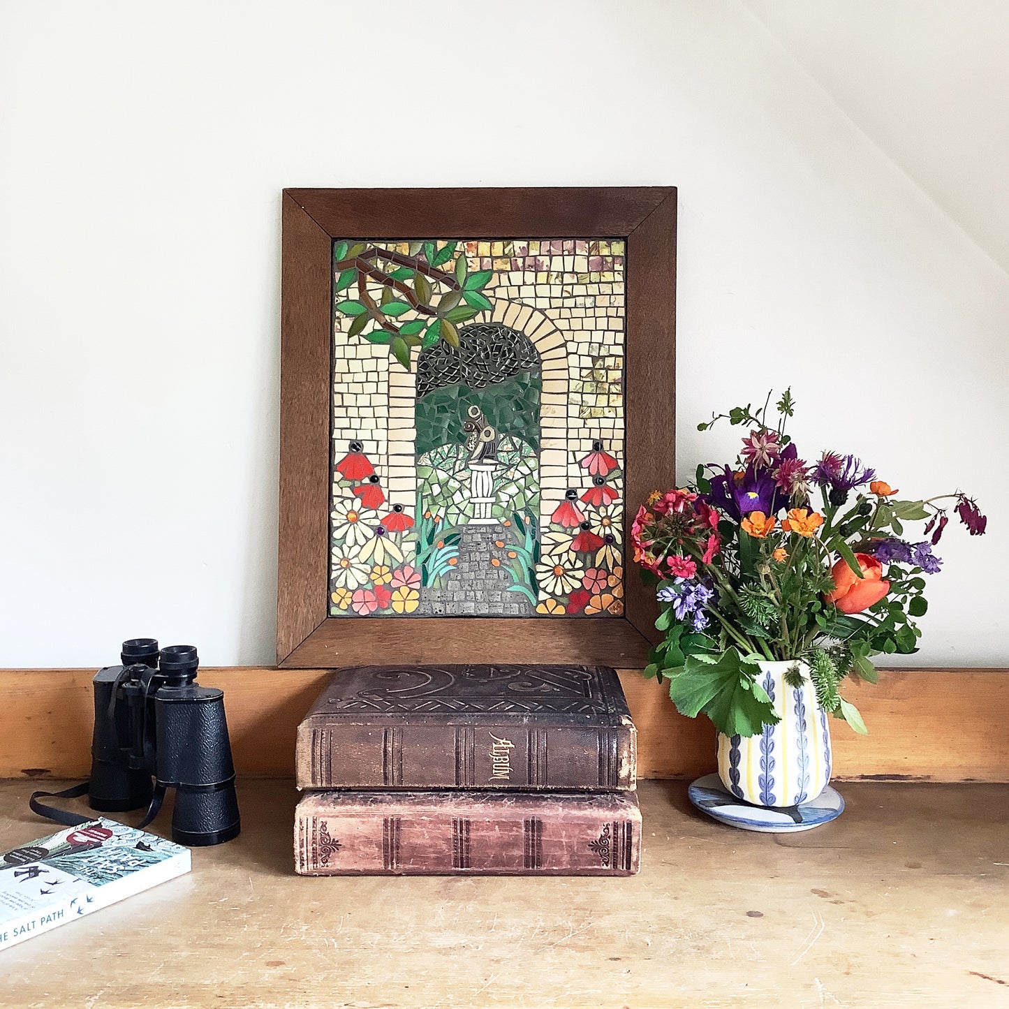 a mosaic of an archway with a branch and flowers, framed in wood, sitting on two books. Flowers and binoculars beside it