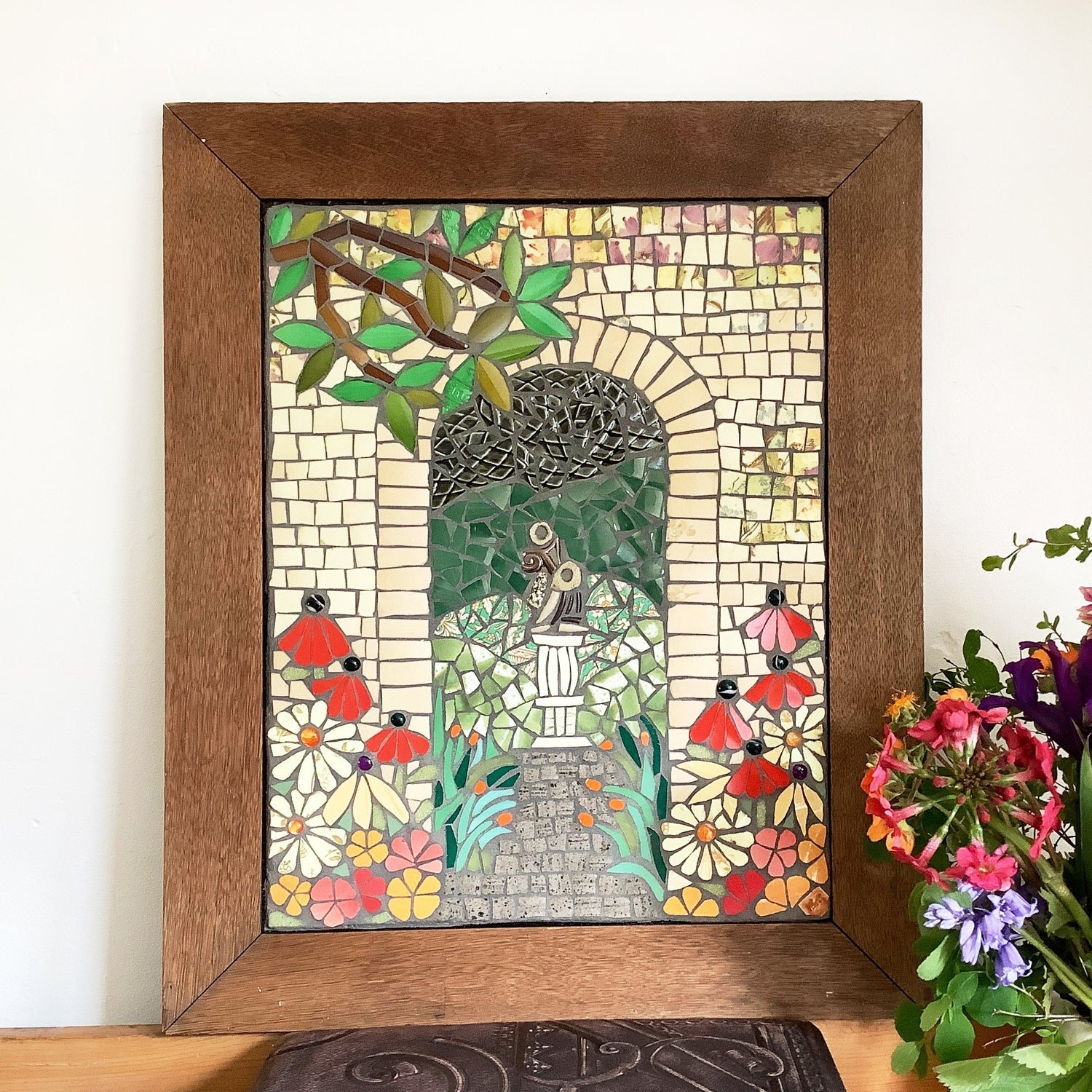 a mosaic of a garden with a featured arch with sculpture. Flowers in the foreground. displayed on a pile of books with flowers beside