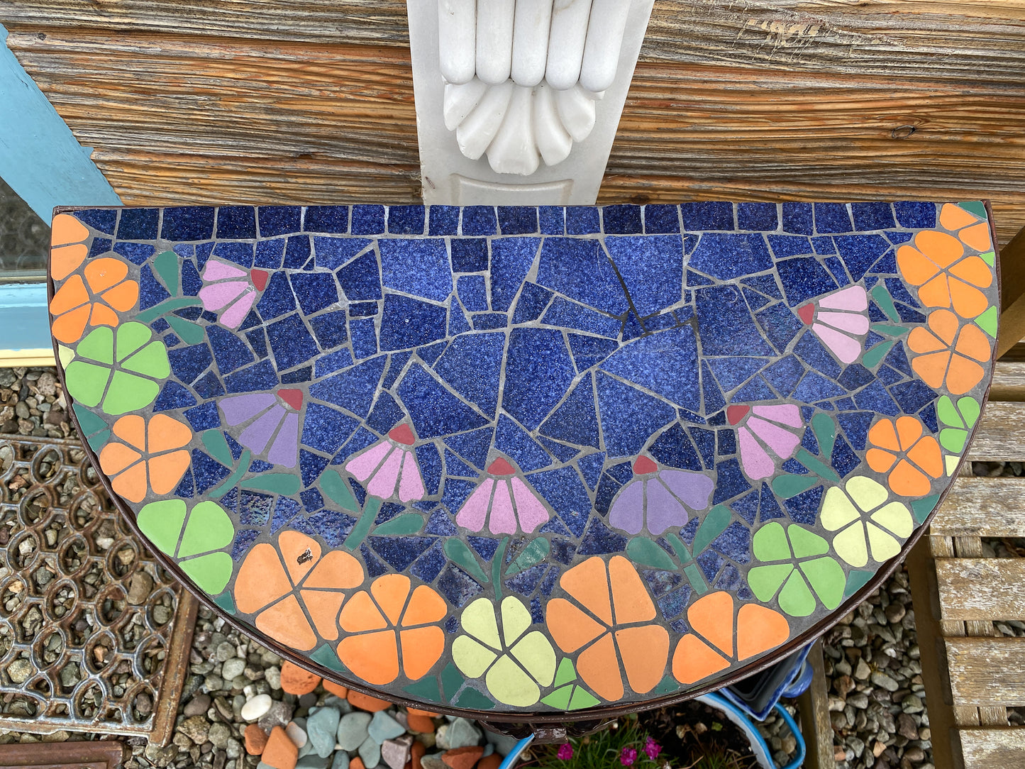 half moon patio table with mosaic surfaces. Orange and lime green flowers edge the blue centre Photographed from above and outdoors