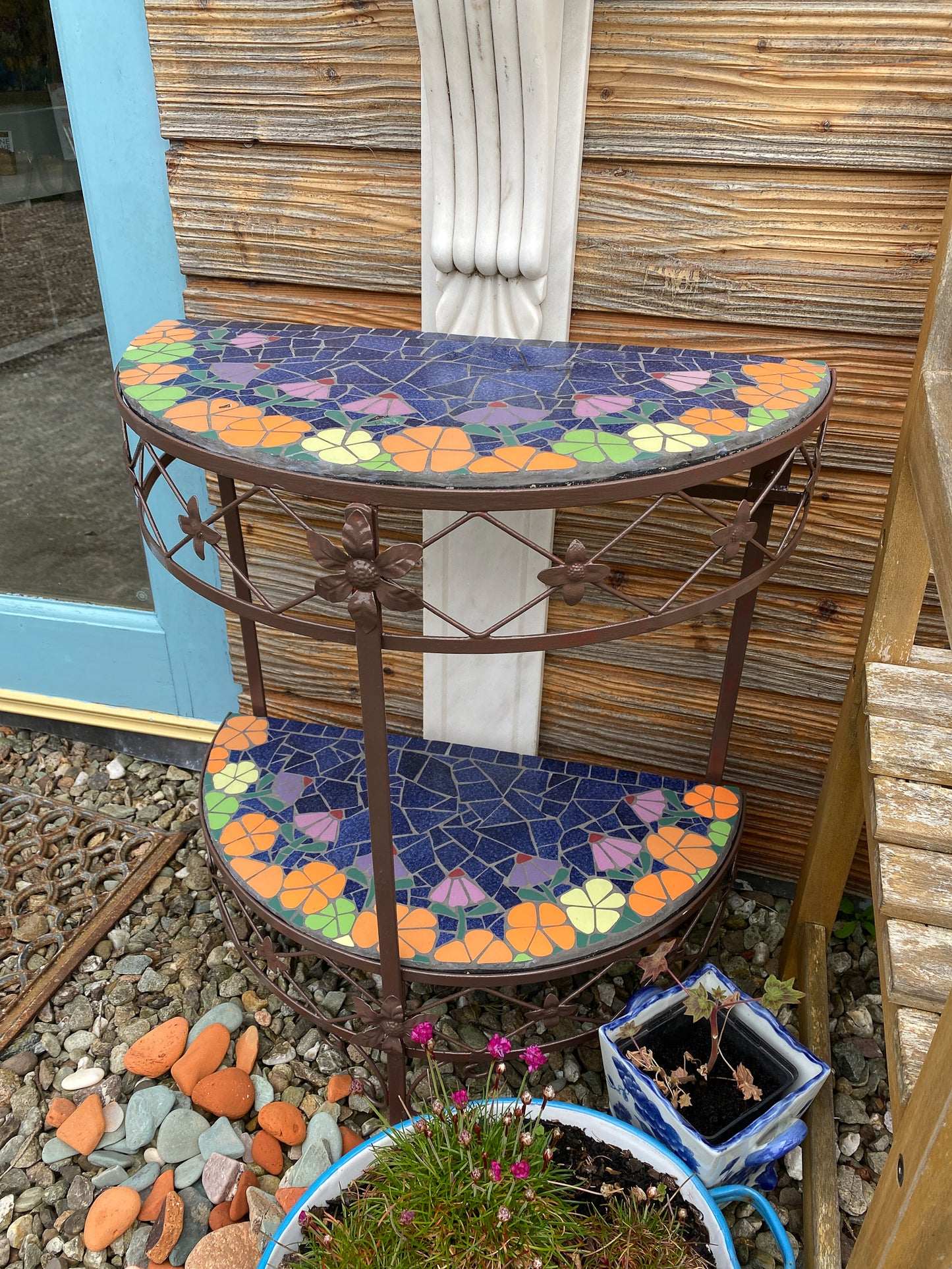 half moon patio table with mosaic surfaces. Two surfaces, with orange and lime green flowers edge the blue centre . The table is a metal vintage one painted dark brown. Photographed straight on and in an outdoors seating area