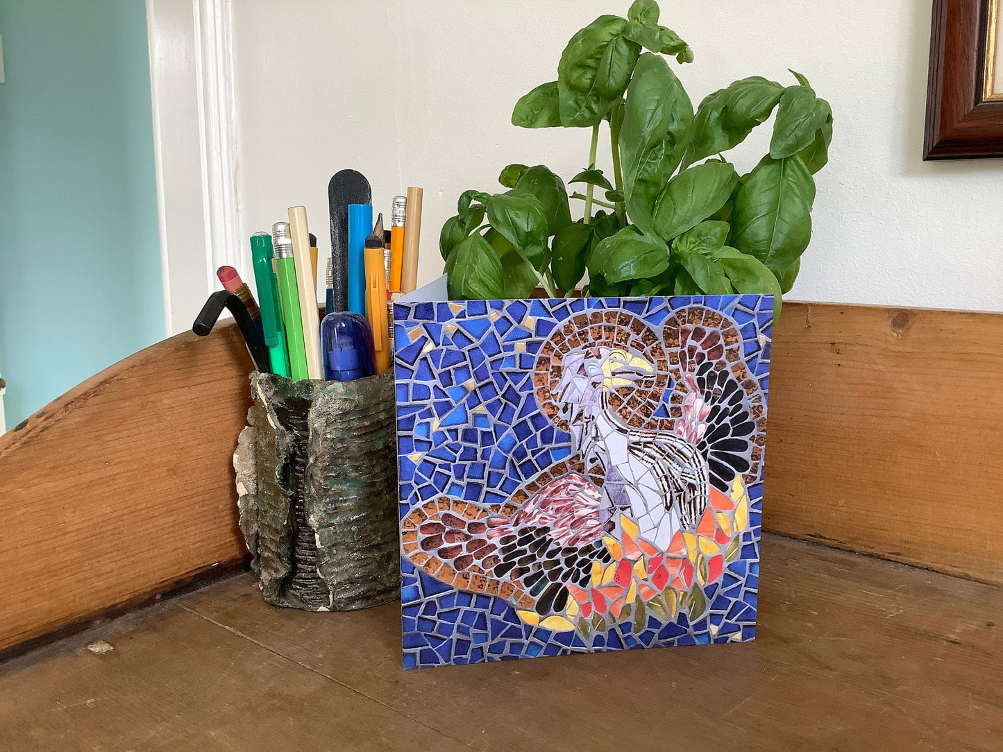 The Phoenix bird rising from the ashes with a halo depicted in mosaic. Dark blue tile and gold background. The bird has a brown body and wings and the fire is in oranges, yellows and red tiles. Photographed on display, alongside a pot of pens and a basil plant on a sideboard