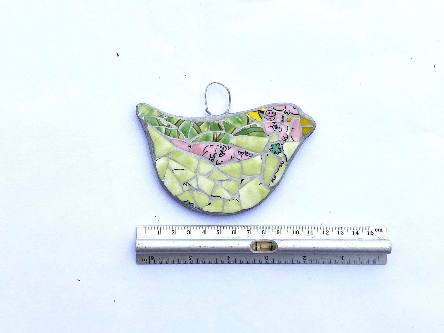 hanging bird mosaic mostly in green bone china, with pink areas showing pigs noses and feet. Shown on a white background with a ruler for scale