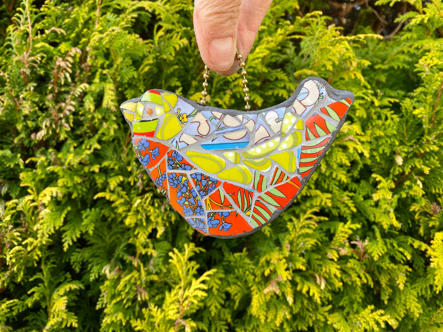 A colourful mosaic bird with red breast, and green head and wings, made from broken crockery. Shown hanging against an evergreen background