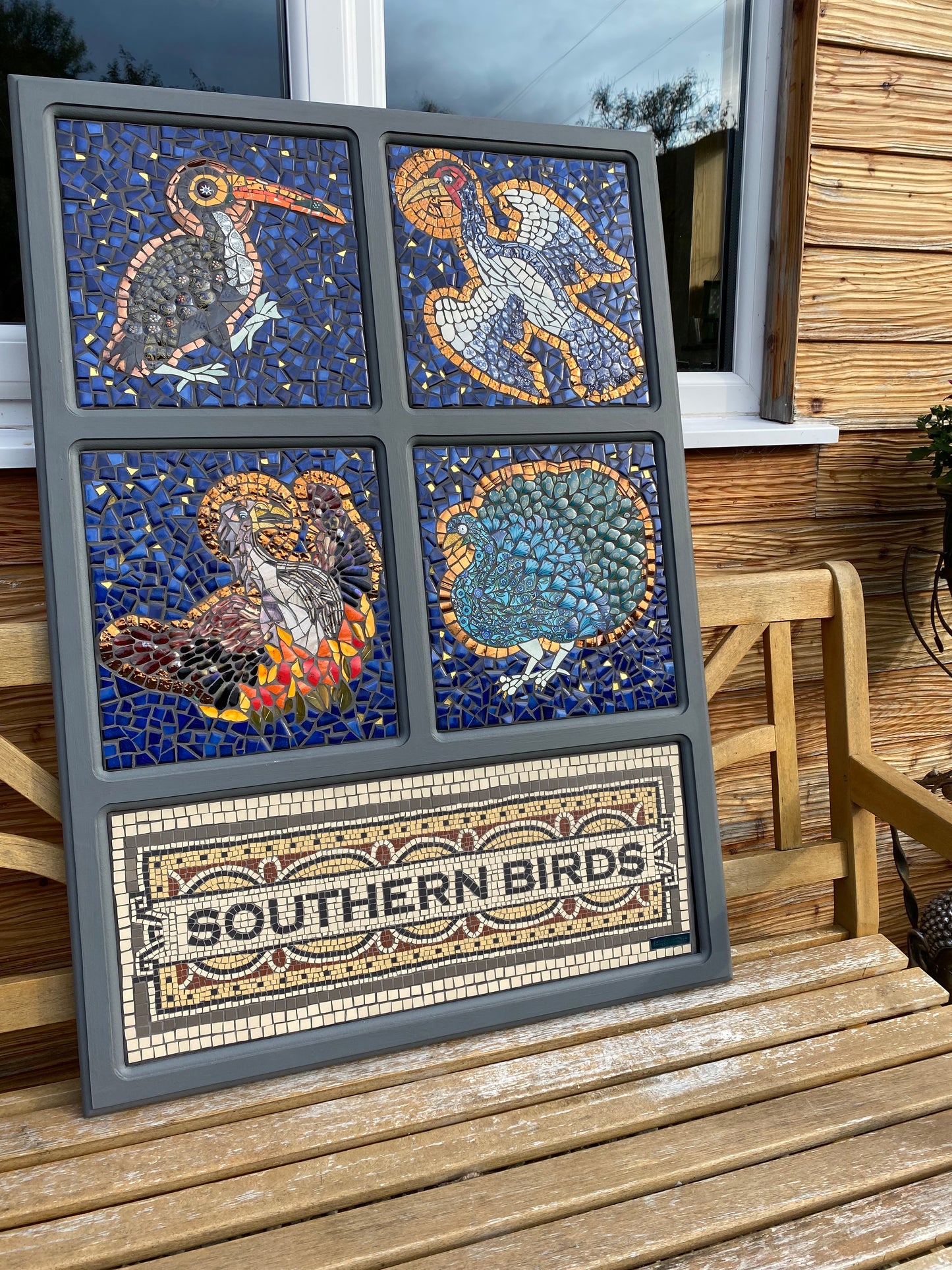 showing the 5-paneled mosaic with four bird panels and one title panel. it is displayed on a bench outside