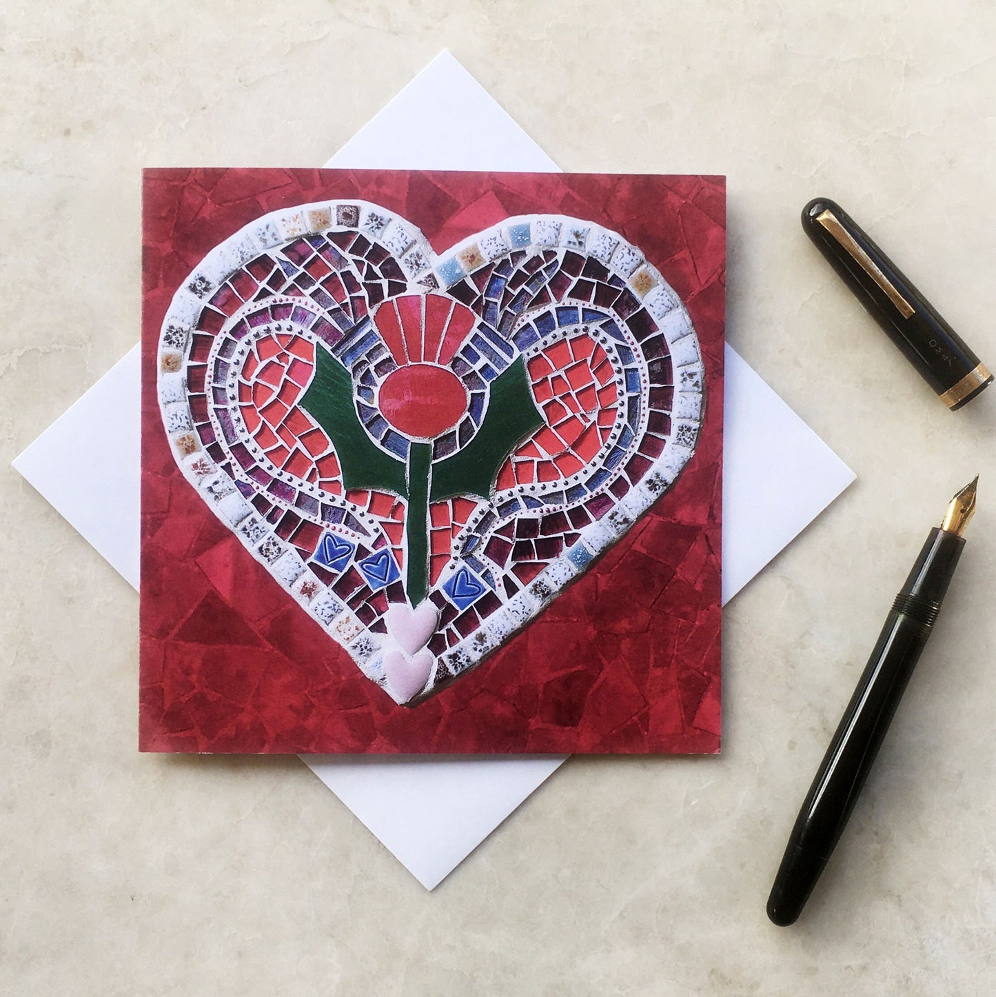 A square card with a thistle on a heart shape, in a mosaic pattern, with a red background. It is photographed with an envelope underneath, on a neutral background. Alongside it is a vintage ink pen.