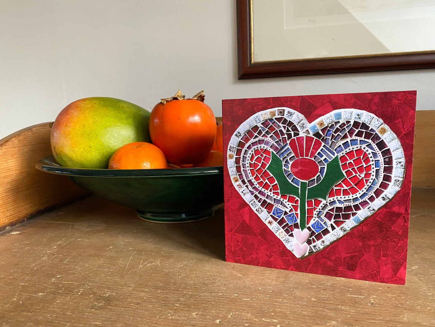 A square card with a thistle on a heart shape, in a mosaic pattern, with a red background. It is photographed on display beside a bowl of fruit on a sideboard