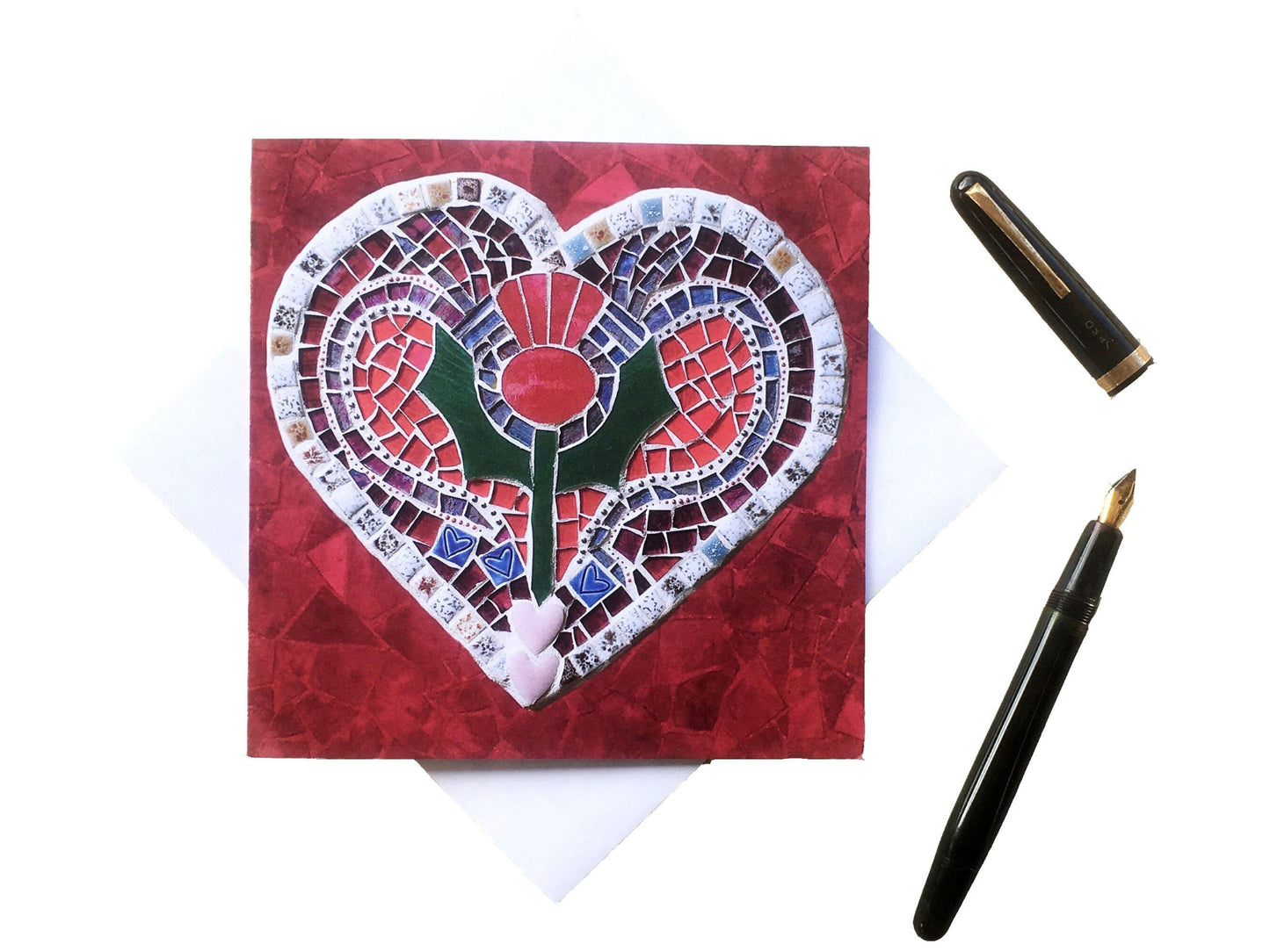A square card with a thistle on a heart shape, in a mosaic pattern, with a red background. It is photographed with an envelope underneath, on a white background. Alongside it is a vintage ink pen.