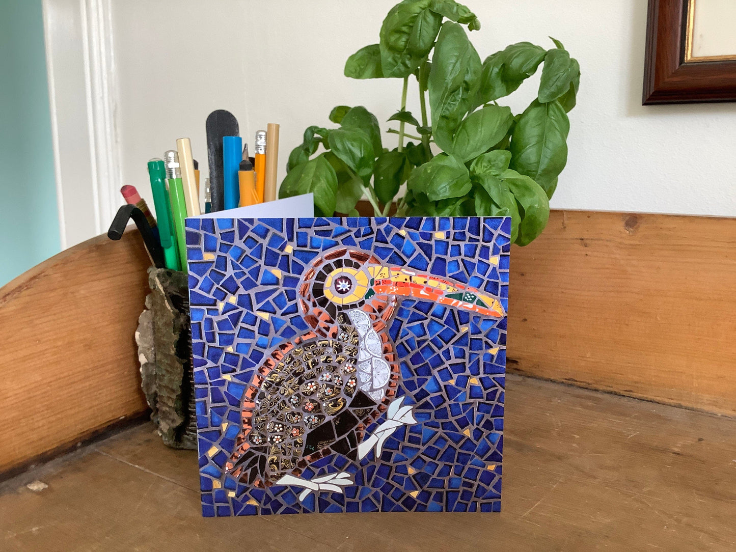Card of a toucan bird with a halo depicted in mosaic. Dark blue tile and gold background. The bird has a black patterned body and a vibrant orange and yellow beak. It has a big orange eye. photographed on a pine sideboard with a pot of pens and a basil plant