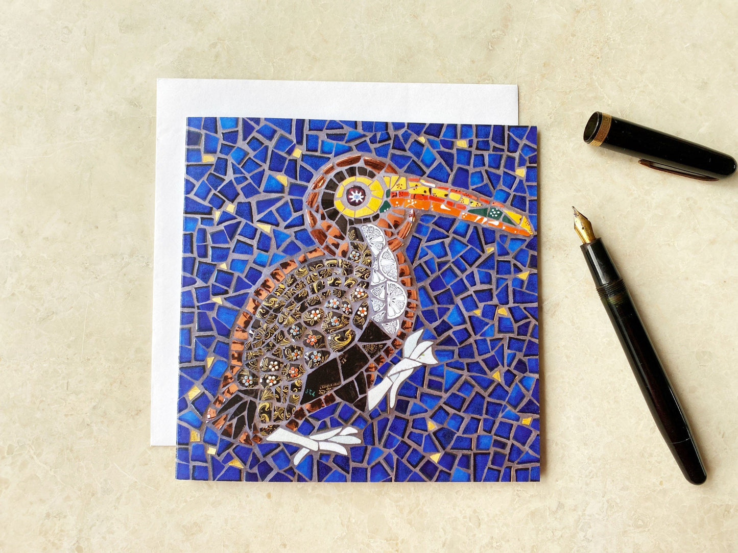 Card of a toucan bird with a halo depicted in mosaic. Dark blue tile and gold background. The bird has a black patterned body and a vibrant orange and yellow beak. It has a big orange eye. photographed on a neutral background with an ink pen alongside