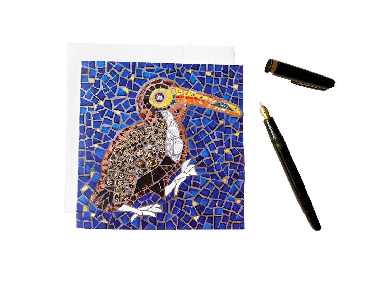 Card of a toucan bird with a halo depicted in mosaic. Dark blue tile and gold background. The bird has a black patterned body and a vibrant orange and yellow beak. It has a big orange eye. photographed on a white background with an ink pen alongside