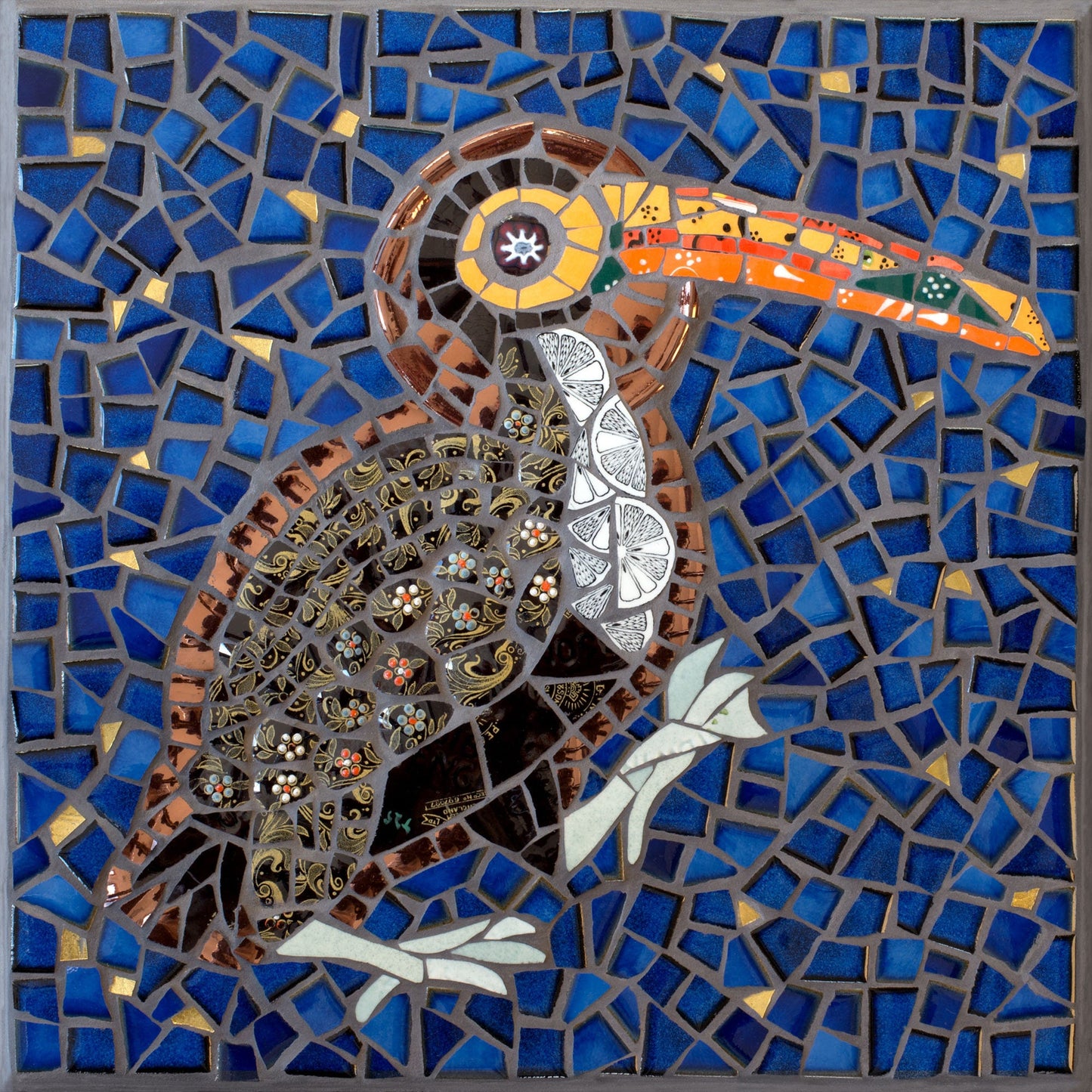 The toucan bird with a halo depicted in mosaic. Dark blue tile and gold background. The bird has a black patterned body and a vibrant orange and yellow beak. Big orange eye