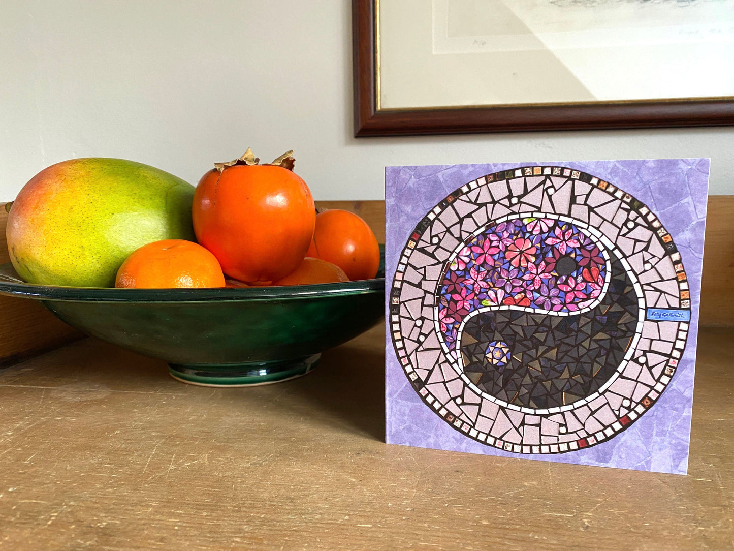 A pink and purple hued mosaic image on a card featurubg a Ying Yang symbol. One half of it is in purple and pink flowers, and the other half is dark grey, with a ribbon of mirror. The ying yang circle is surrounded by purple, for this square card. photographed alongside a bowl of fruit on a sideboard, as if it has just been received