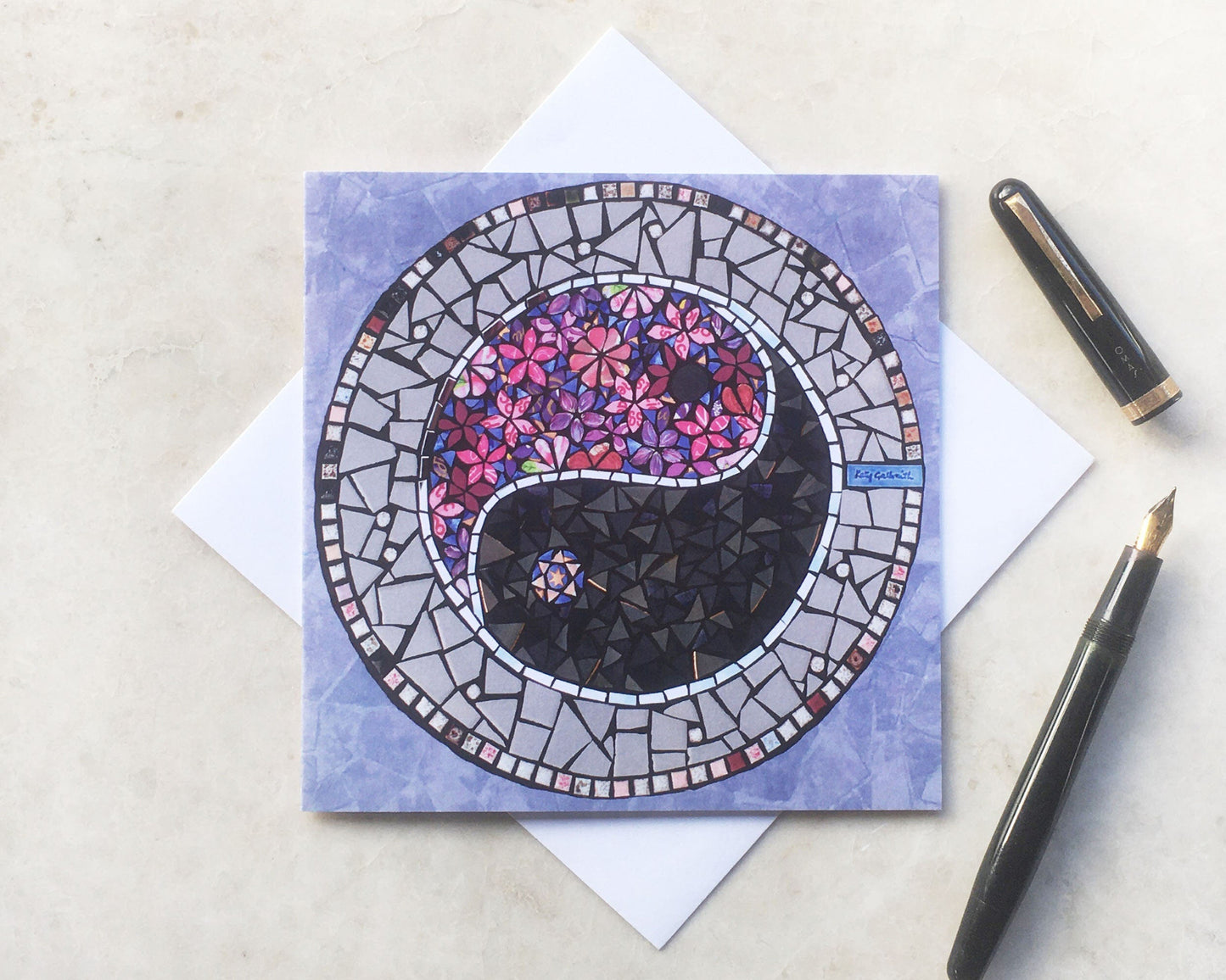 A pink and purple hued mosaic with a Ying Yang symbol. One half of it is in purple and pink flowers, and the other half is dark grey, with a ribbon of mirror. The ying yang circle is surrounded by purple, for this square card. photographed with an ink pen and envelope on a neutral background