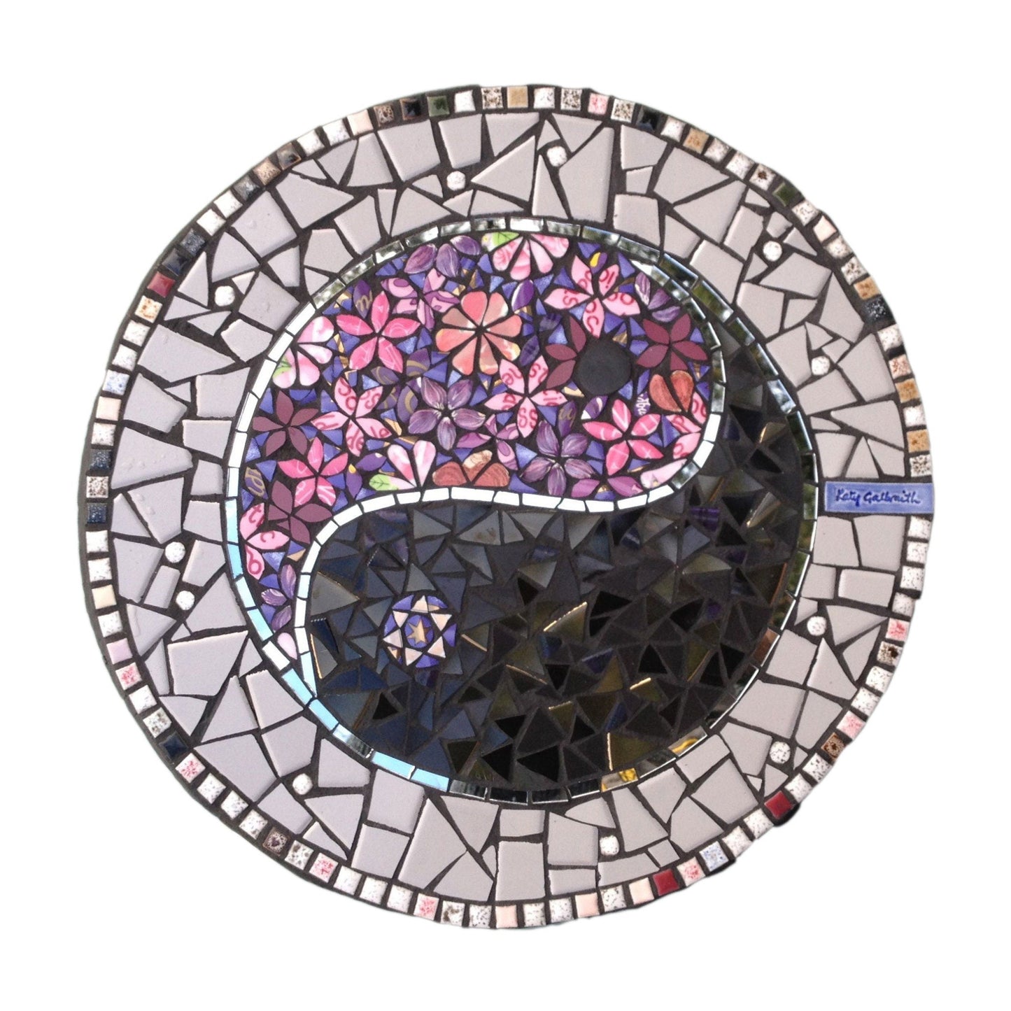 A pink and purple hued mosaic stepping stone with a Ying Yang symbol. One half of it is in purple and pink flowers, and the other half is dark grey, with a ribbon of mirror. Photographed with a white background