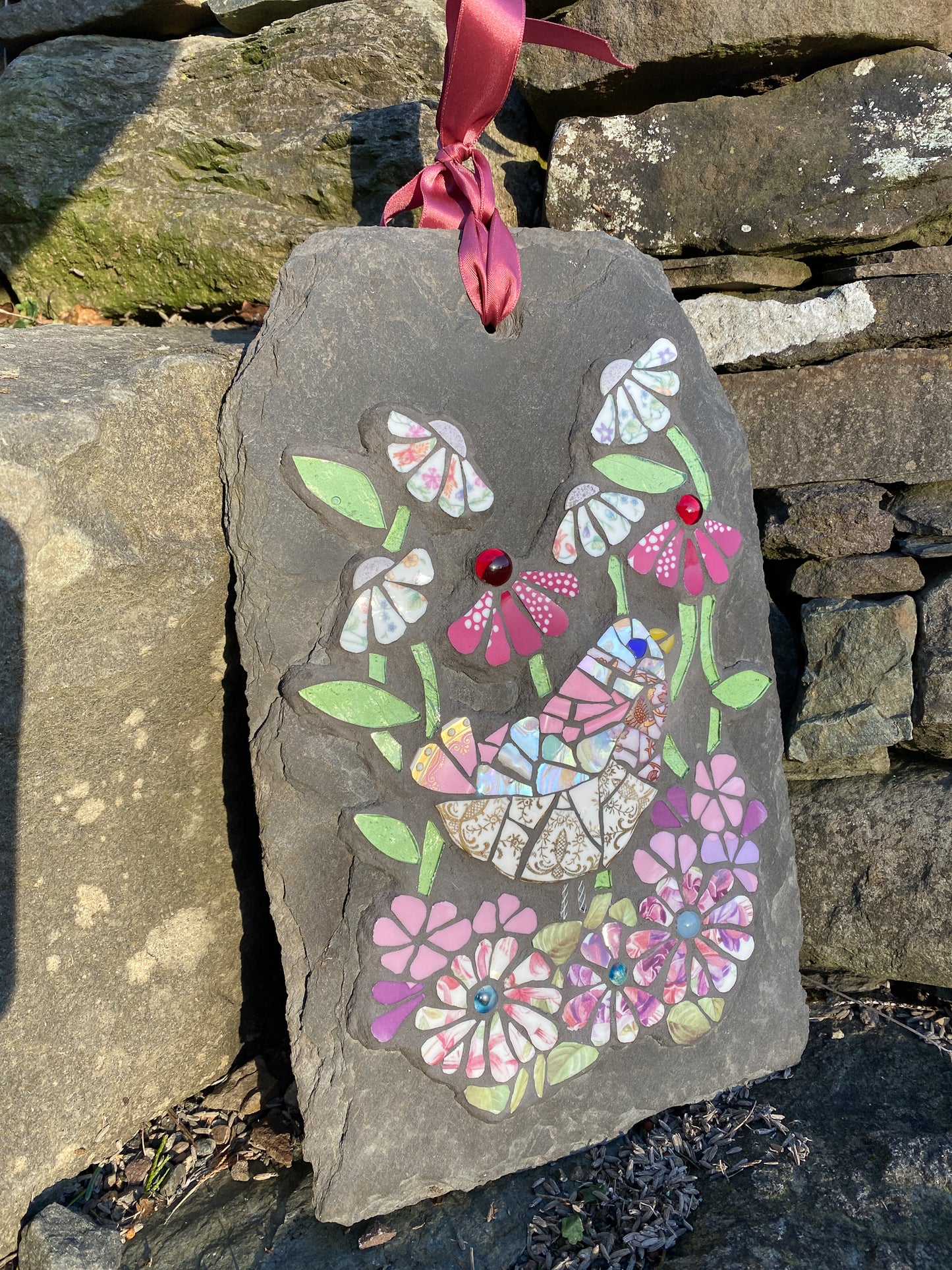 mosaic on slate of a pink gold and white bird surrounded by mosaic flowers in pink, hot pink and chintz tones. leaves in pale green glass. Shown on a stone step