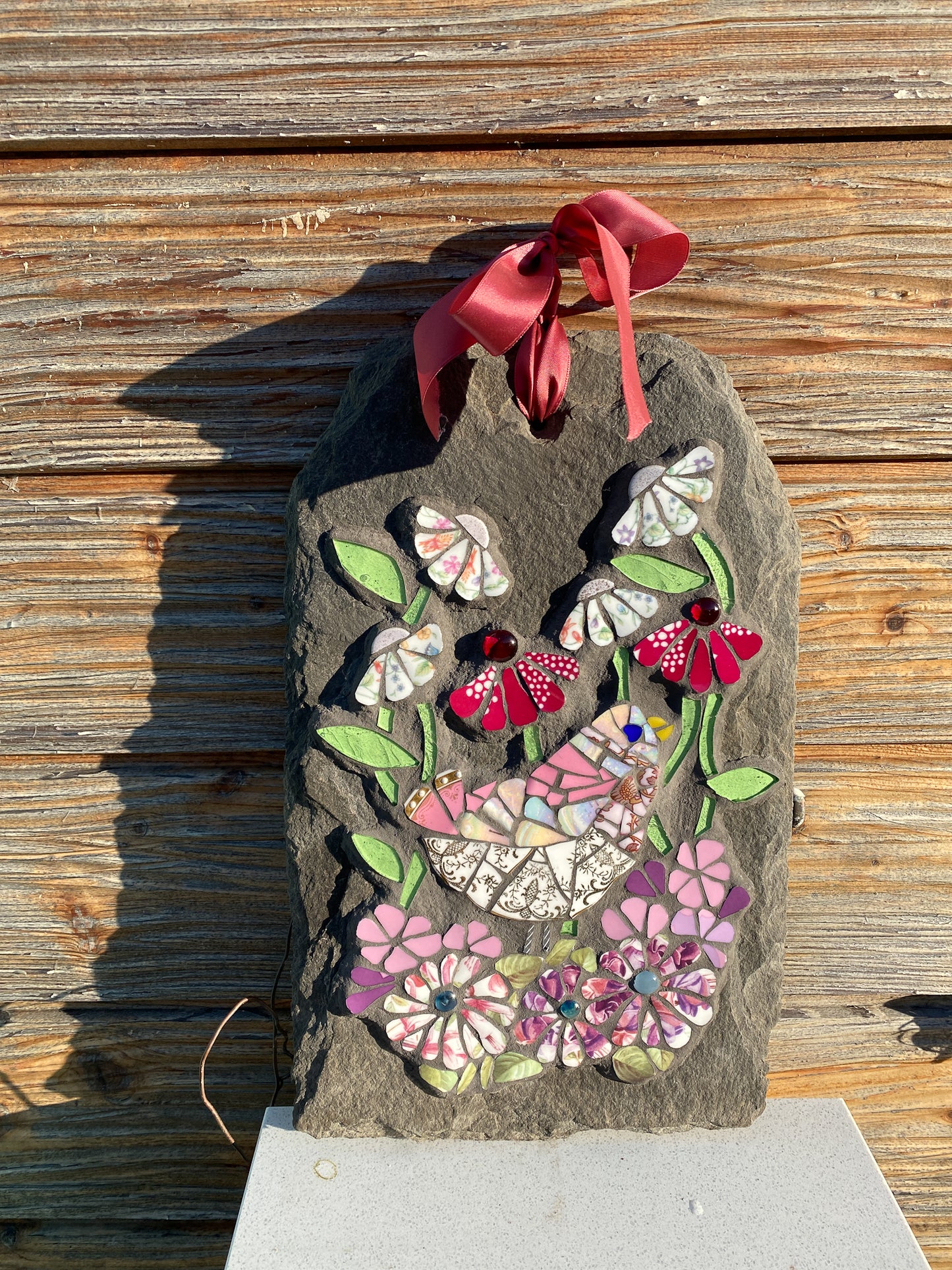 mosaic on slate of a pink gold and white bird surrounded by mosaic flowers in pink, hot pink and chintz tones. leaves in pale green glass. Shown against a wooden background