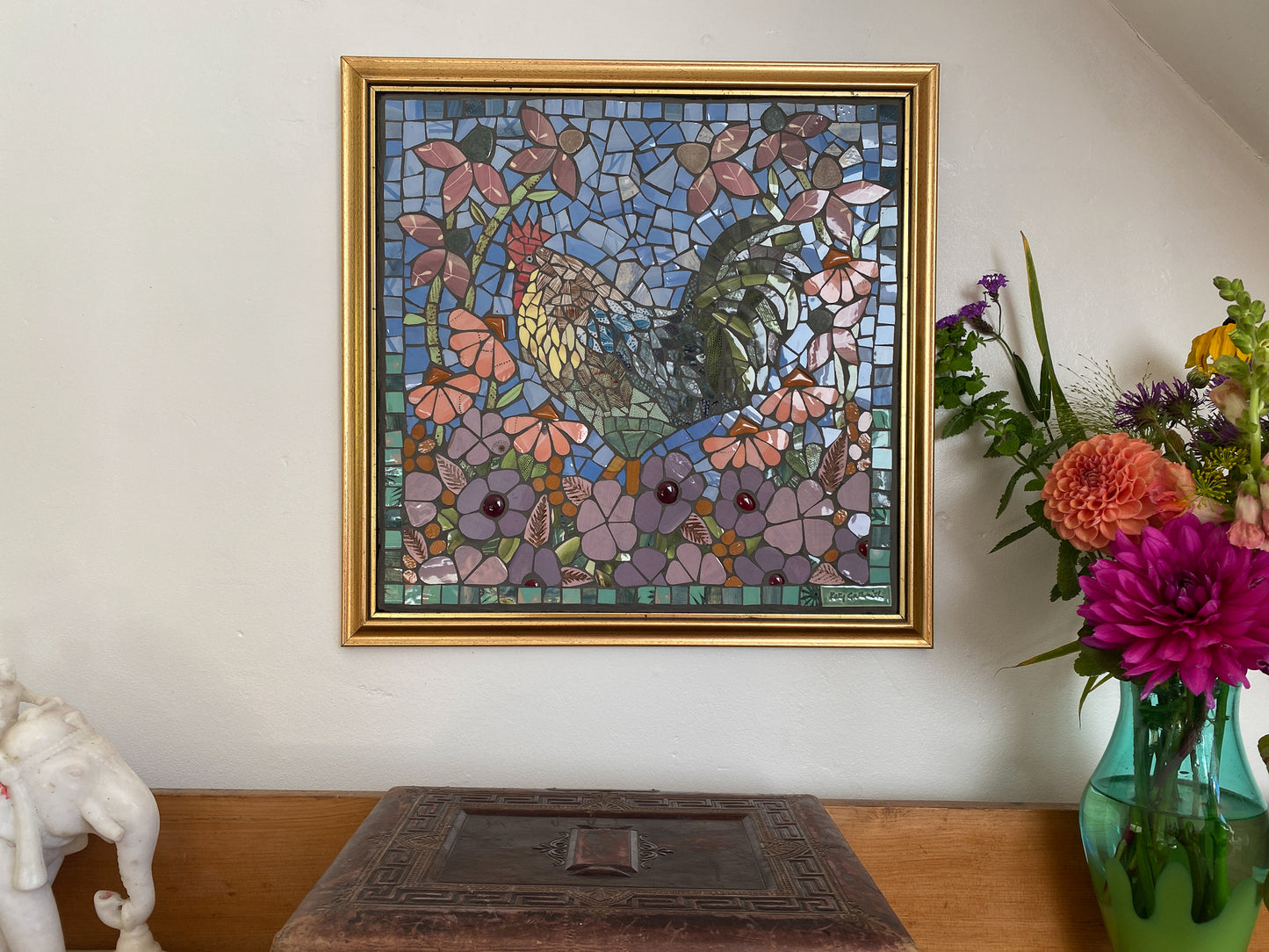 Framed mosaic artwork of flowers and leaves on a wall above a table with a vase of flowers.