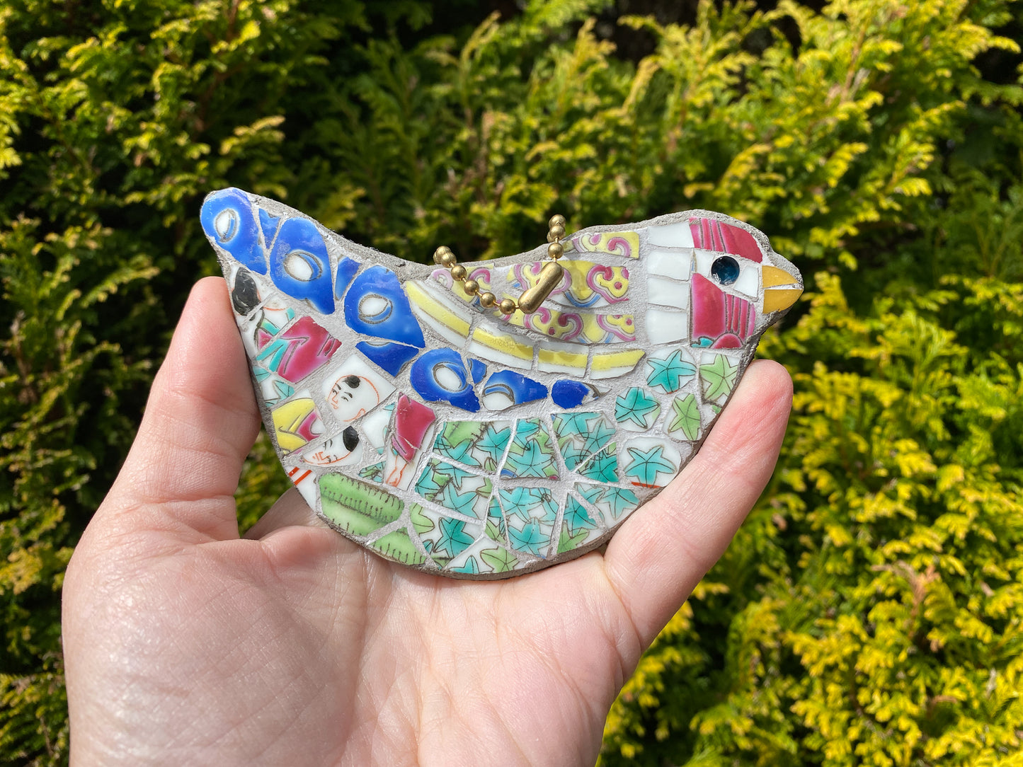a multicoloured bird mosaic which hangs from a ball chain loop, with a red head, green body and blue feathers, all made from a chinese porcelain pot, including some faces. photographed being held in a hand to show size and against a foliage background