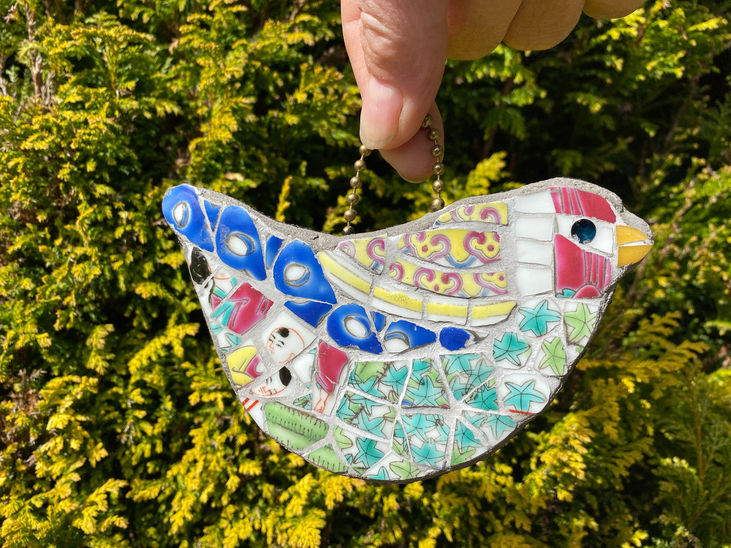 a multicoloured bird mosaic which hangs from a ball chain loop, with a red head, green body and blue feathers, all made from a chinese porcelain pot. photographed hanging by the ball chain and against a foliage background