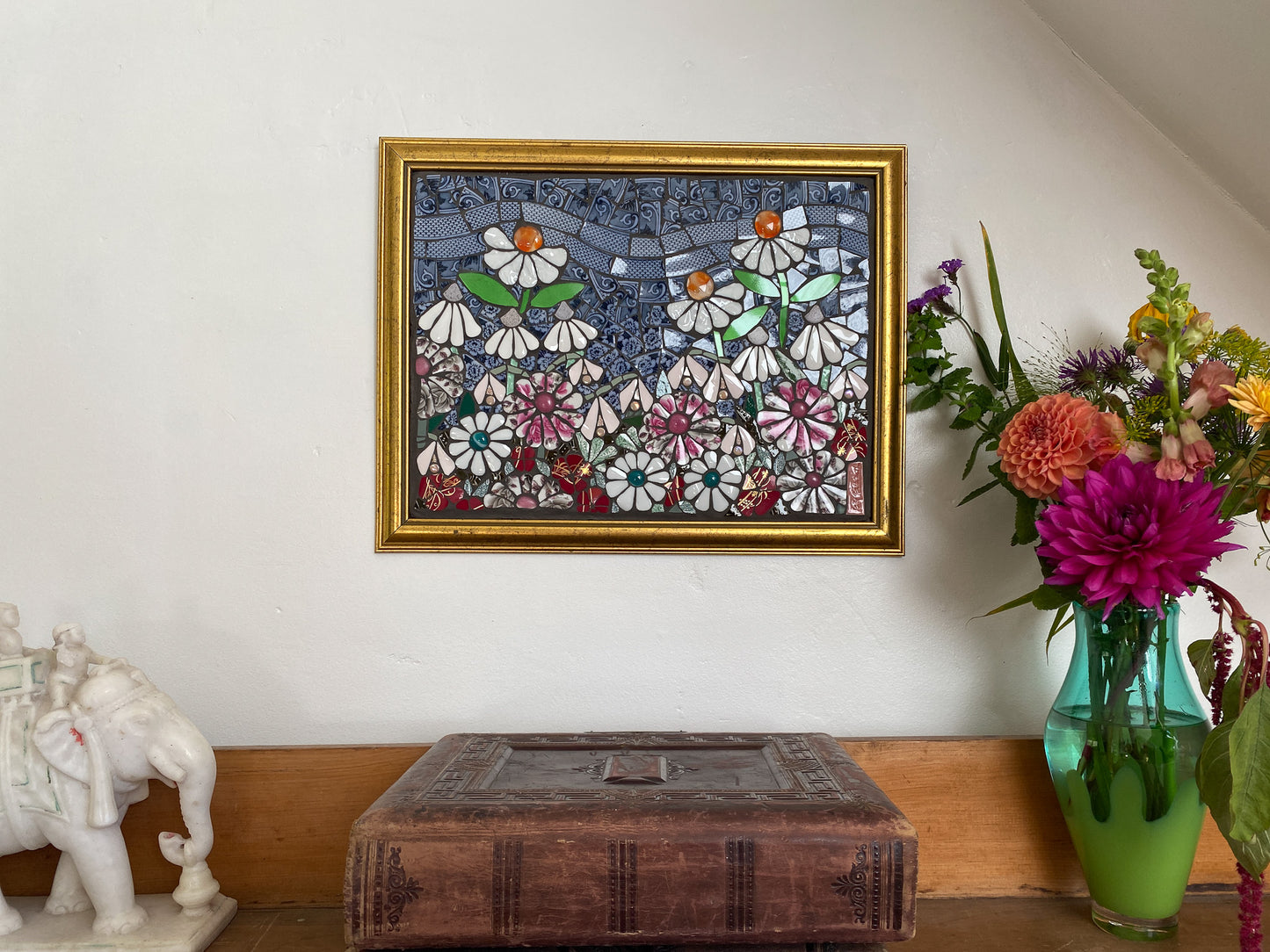 Framed mosaic artwork with flowers on a wall next to a vase of flowers and decorative items.