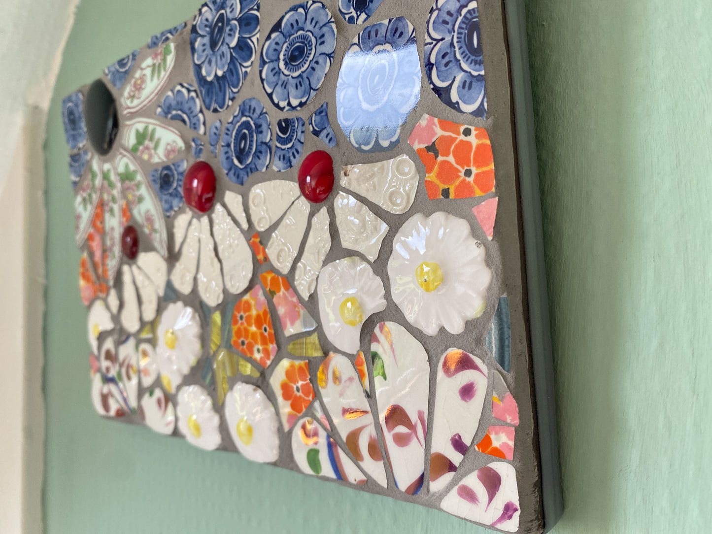 small mosaic flower garden, with white; white and patterned, orange petals, and a blue sky with cut outs of crockery. Red and dark purple glass nuggets. Photographed at an angle, showing the lead strip on the edge