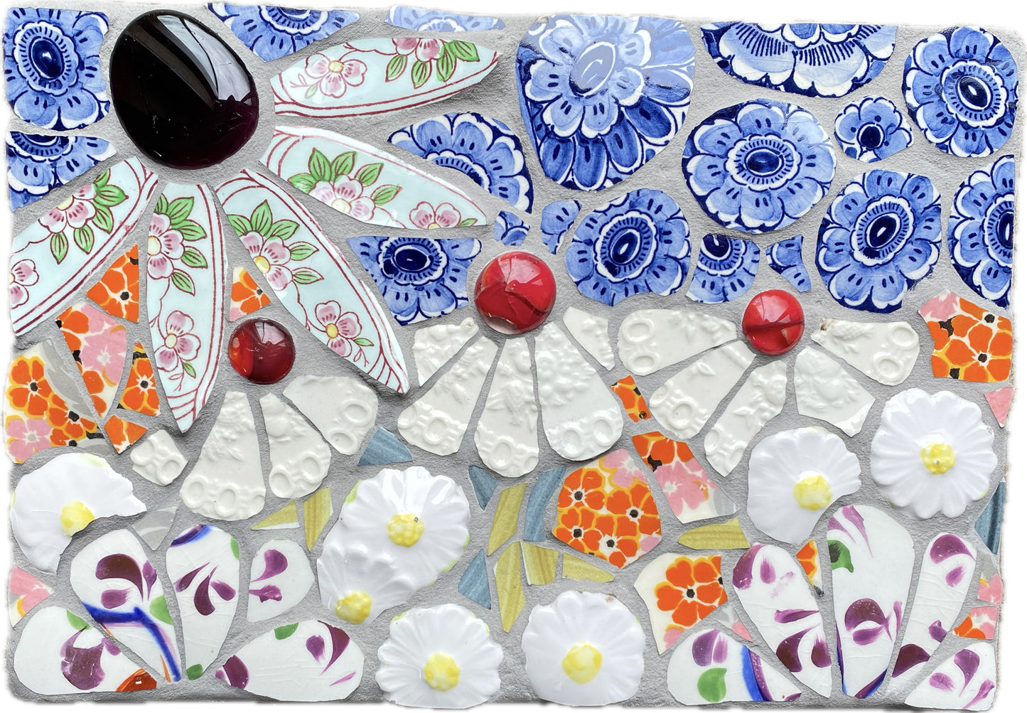 small mosaic flower garden, with white; white and patterned, orange petals, and a blue sky with cut outs of crockery. Red and dark purple glass nuggets.